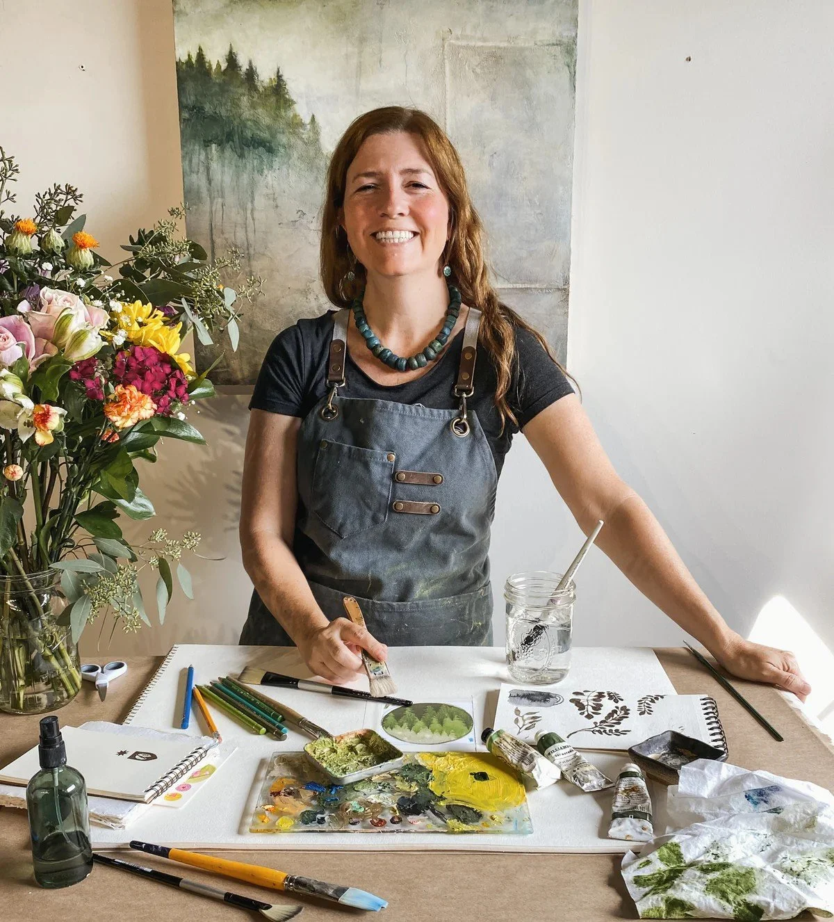 A woman with long brown hair smiling while standing at a table with art supplies, including paint brushes, paints, and sketchbooks. She is wearing a black t-shirt, an apron, and a beaded necklace. There is a large bouquet of colorful flowers on her left side and a painting of a landscape in the background.