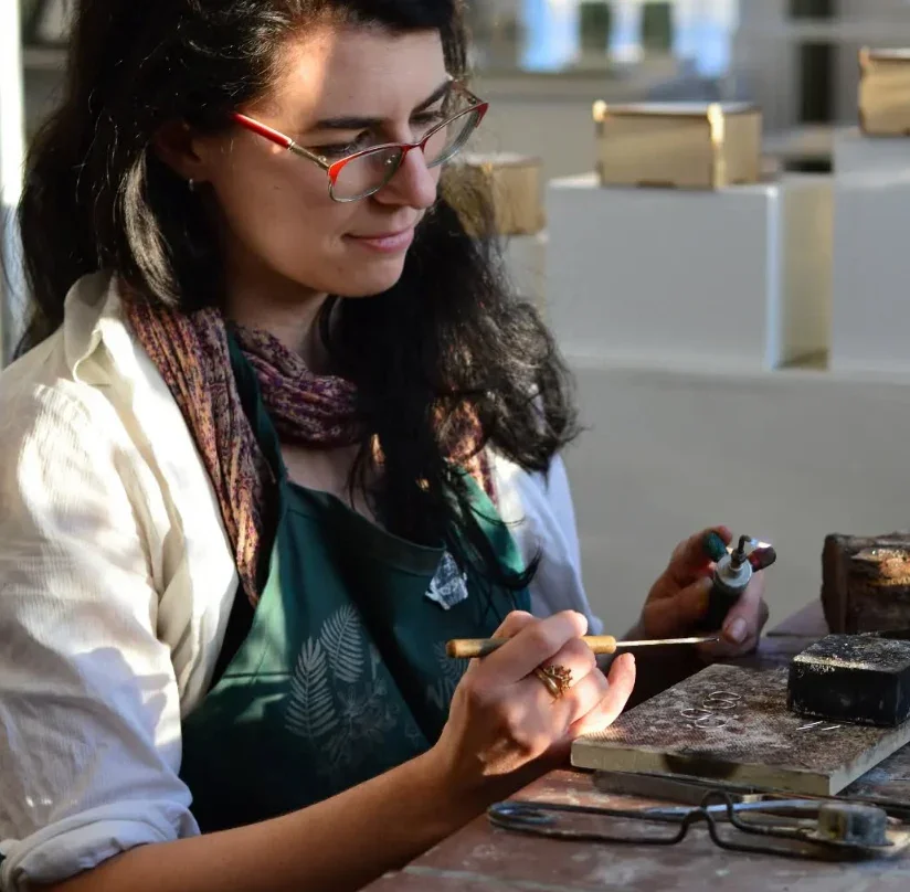 Woman with glasses and dark hair working on jewelry with tools at a workbench.