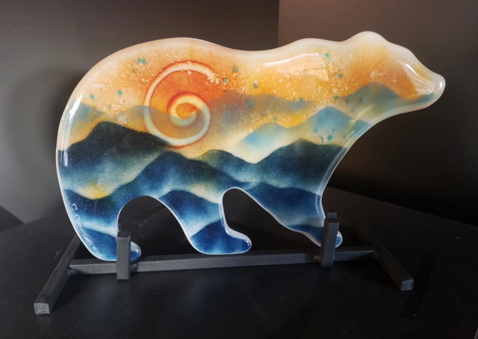 Decorative glass art piece depicting a bear with a landscape of mountains, sun, and sky painted inside, displayed on a black stand.