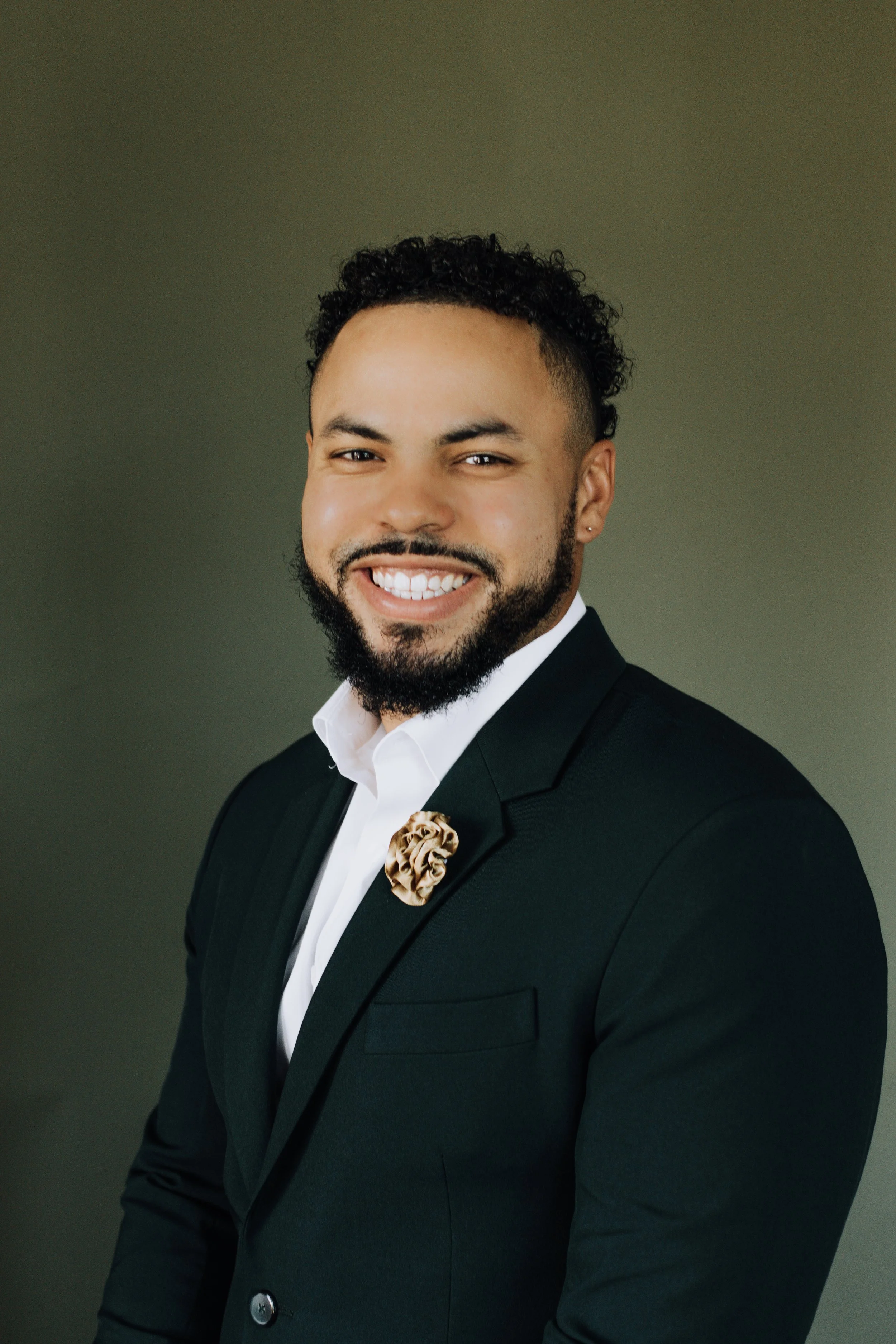 Portrait of a smiling man in a black suit with a decorative pin, white shirt, and a neutral background.