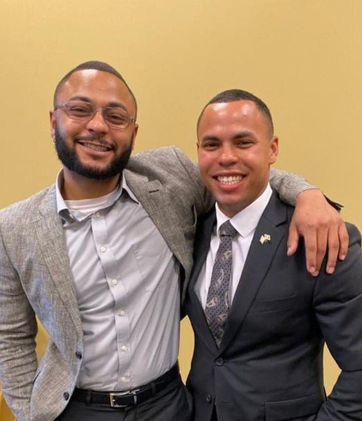 Two men smiling with their arms around each other, dressed in business attire, standing against a yellow background.