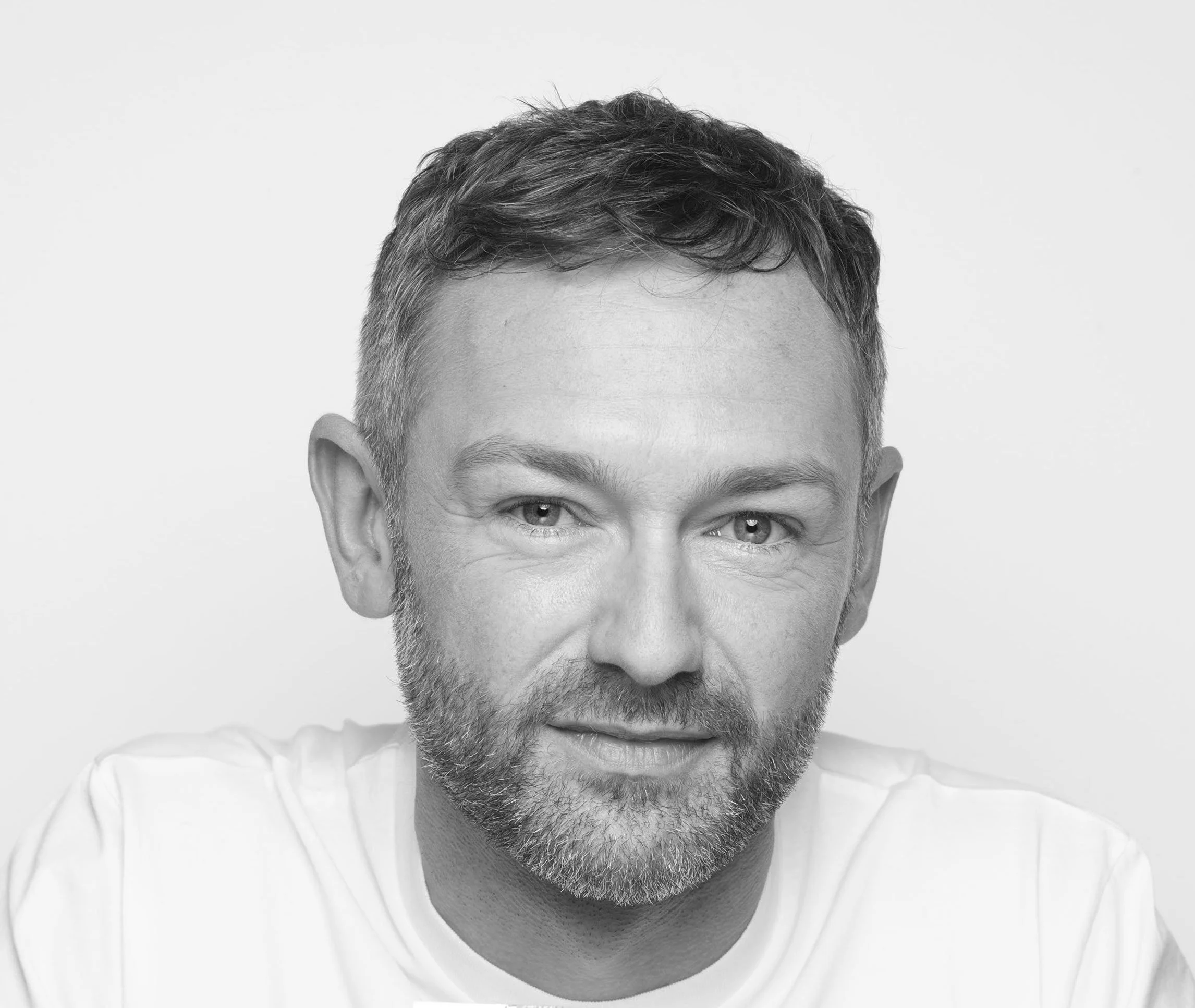 Black-and-white portrait of a man with short wavy hair and a beard, smiling slightly, wearing a white shirt, against a plain light background.