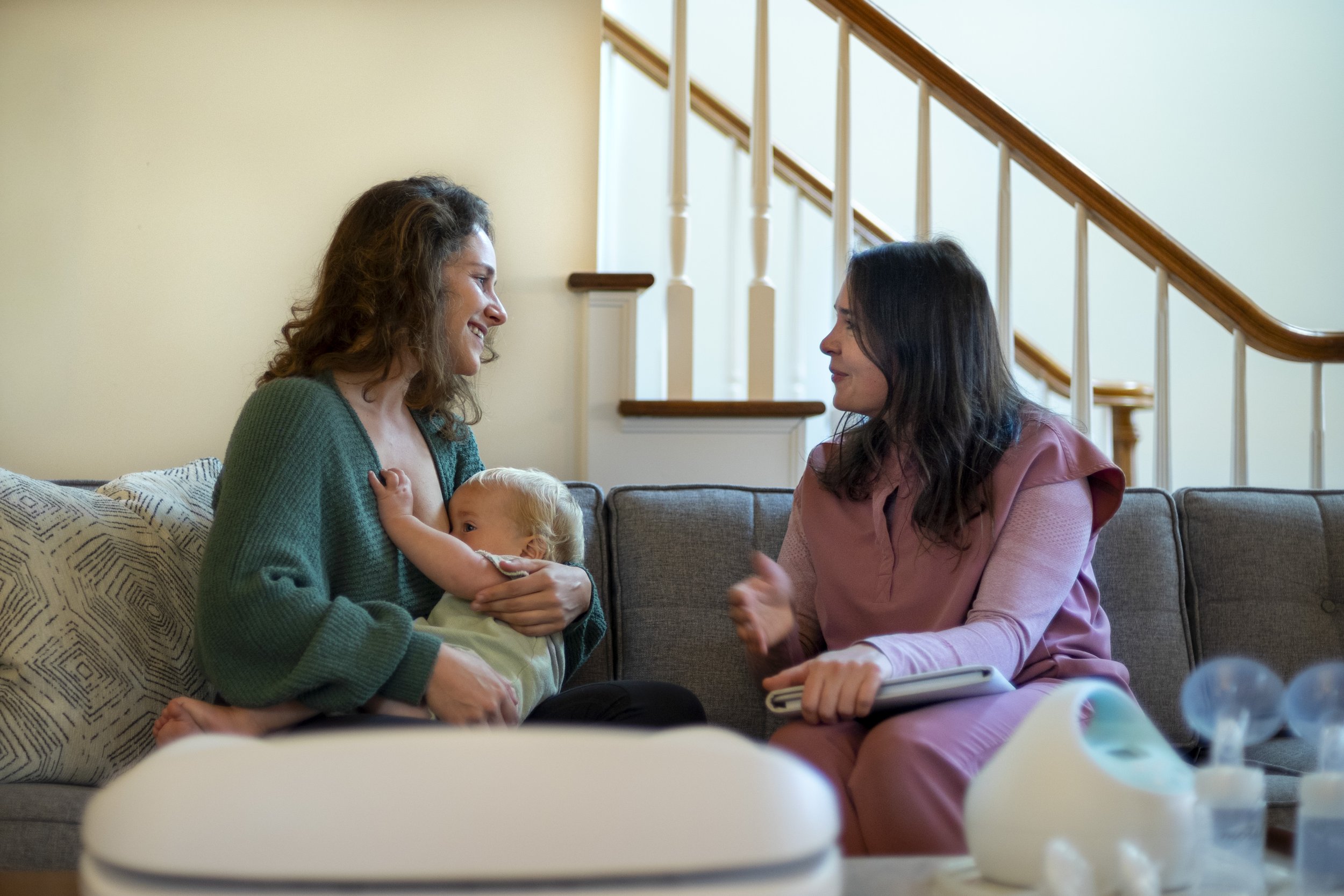 Two women talking on a gray couch with a baby and a woman holding a laptop and a baby