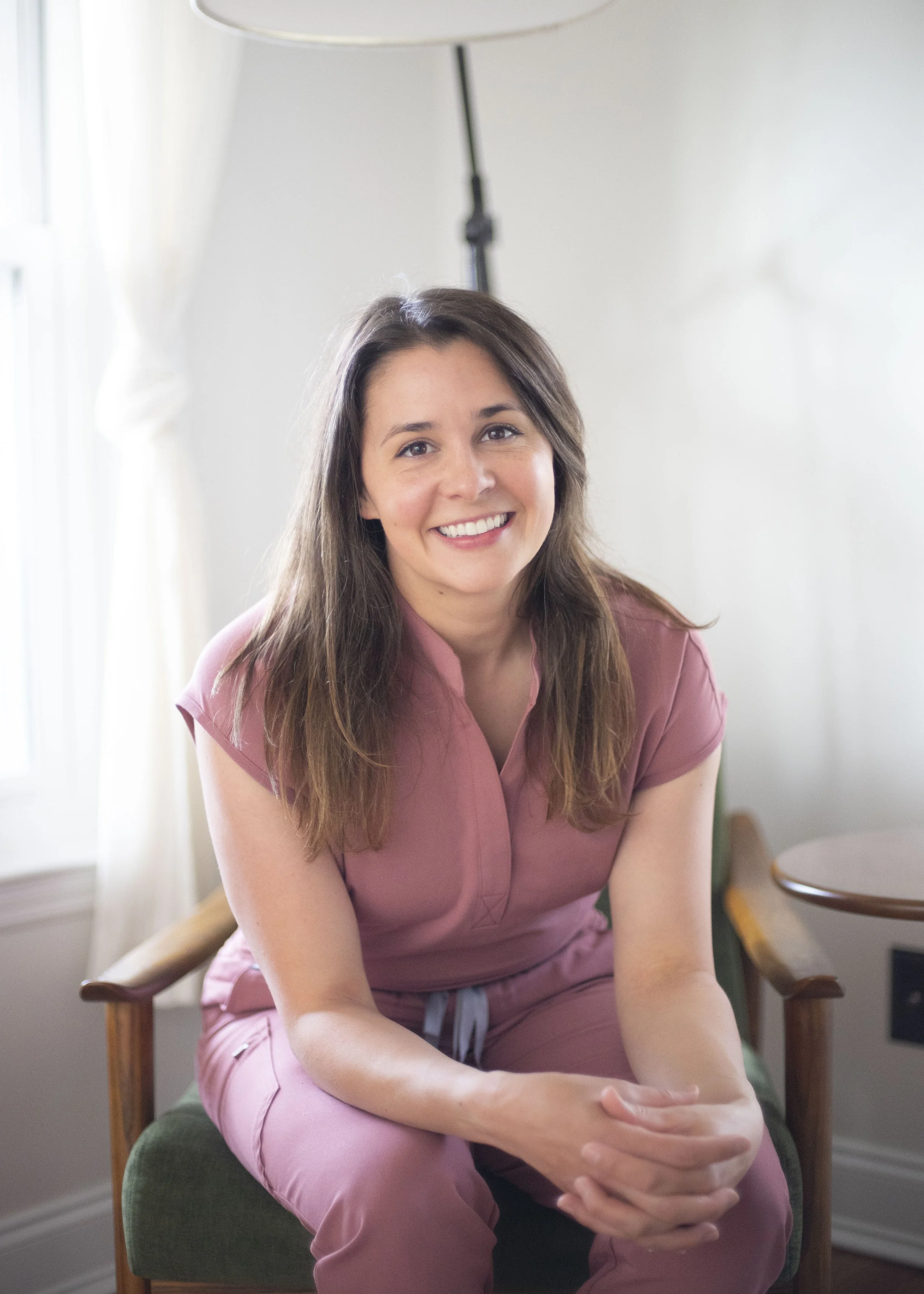 A smiling woman with long brown hair wearing pink scrubs sitting on a green chair in a well-lit room.