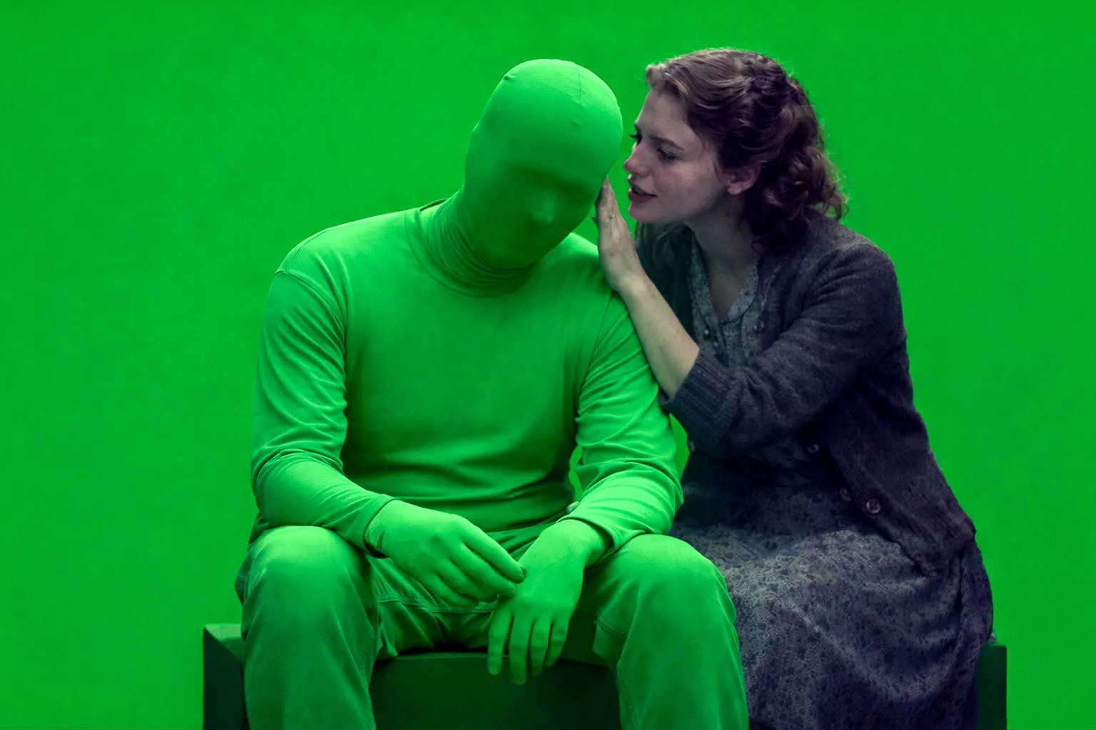 A woman leaning close to a person dressed in a full green suit with a matching green face mask, sitting on a bench against a green screen background.