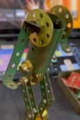 A green and yellow mechanical or robotic arm with a rounded, open-end design, possibly a toy or a model, in a room with shelves in the background.