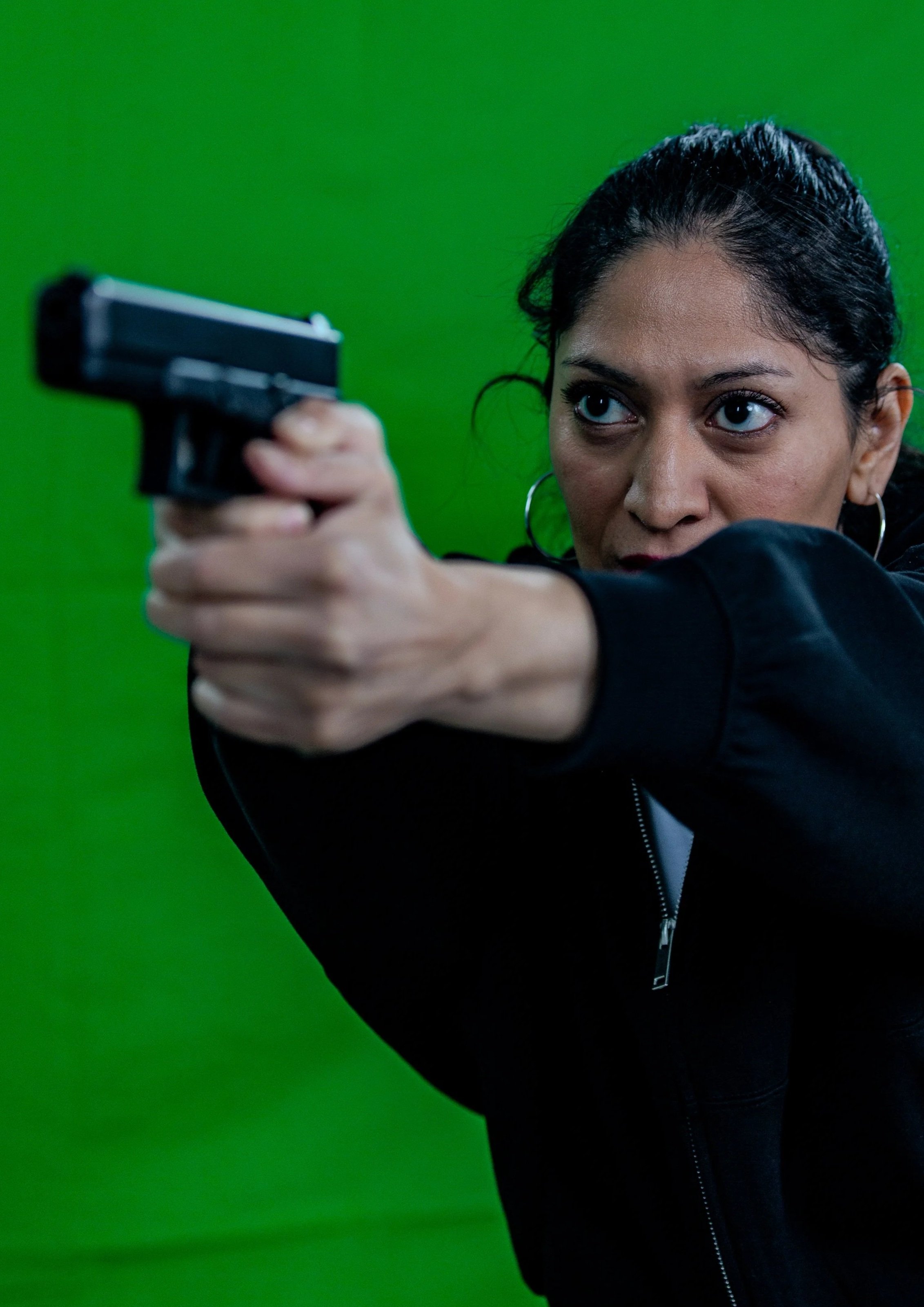 Isabel Rainer Heart with dark hair and hoop earrings aiming a black handgun against a green background.