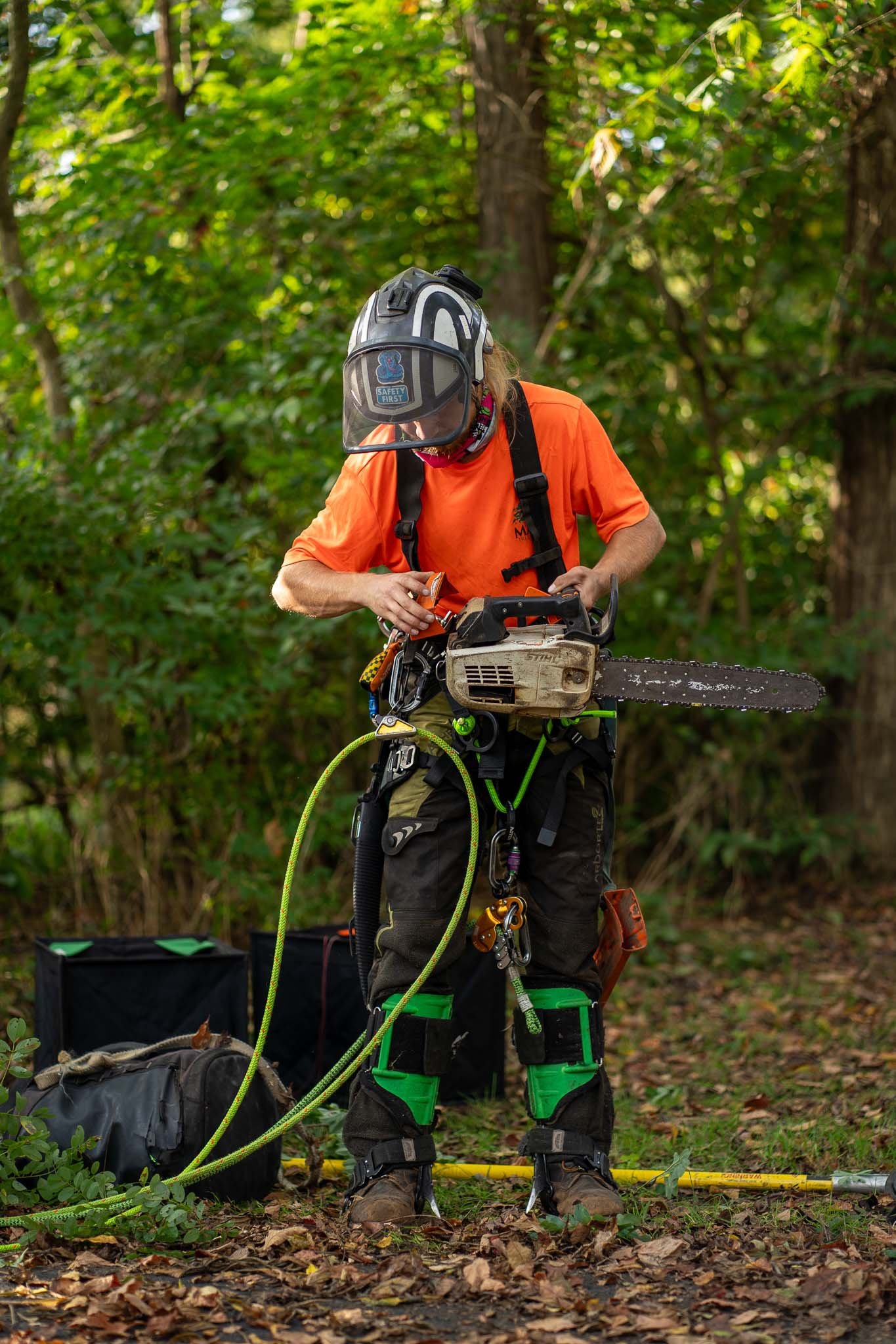 A person wearing safety gear and protective clothing, including a helmet and harness, stands outdoors in a wooded area, preparing equipment, with tools and gear on the ground.