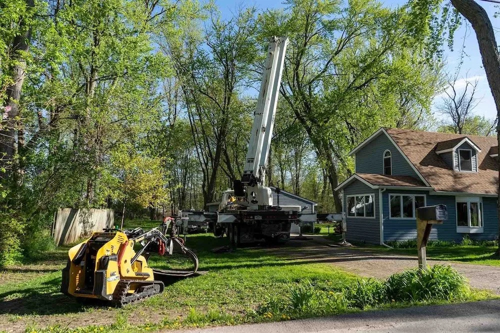 Residential area with tree removal equipment including a crane and mini loader on a grassy yard near a blue house.