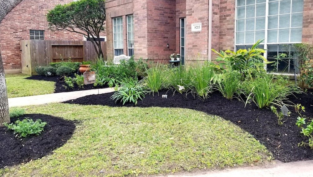 Front yard with landscaped garden, including shrubs, potted plants, green lawn, and mulch around plants, near a brick house and wooden fence.