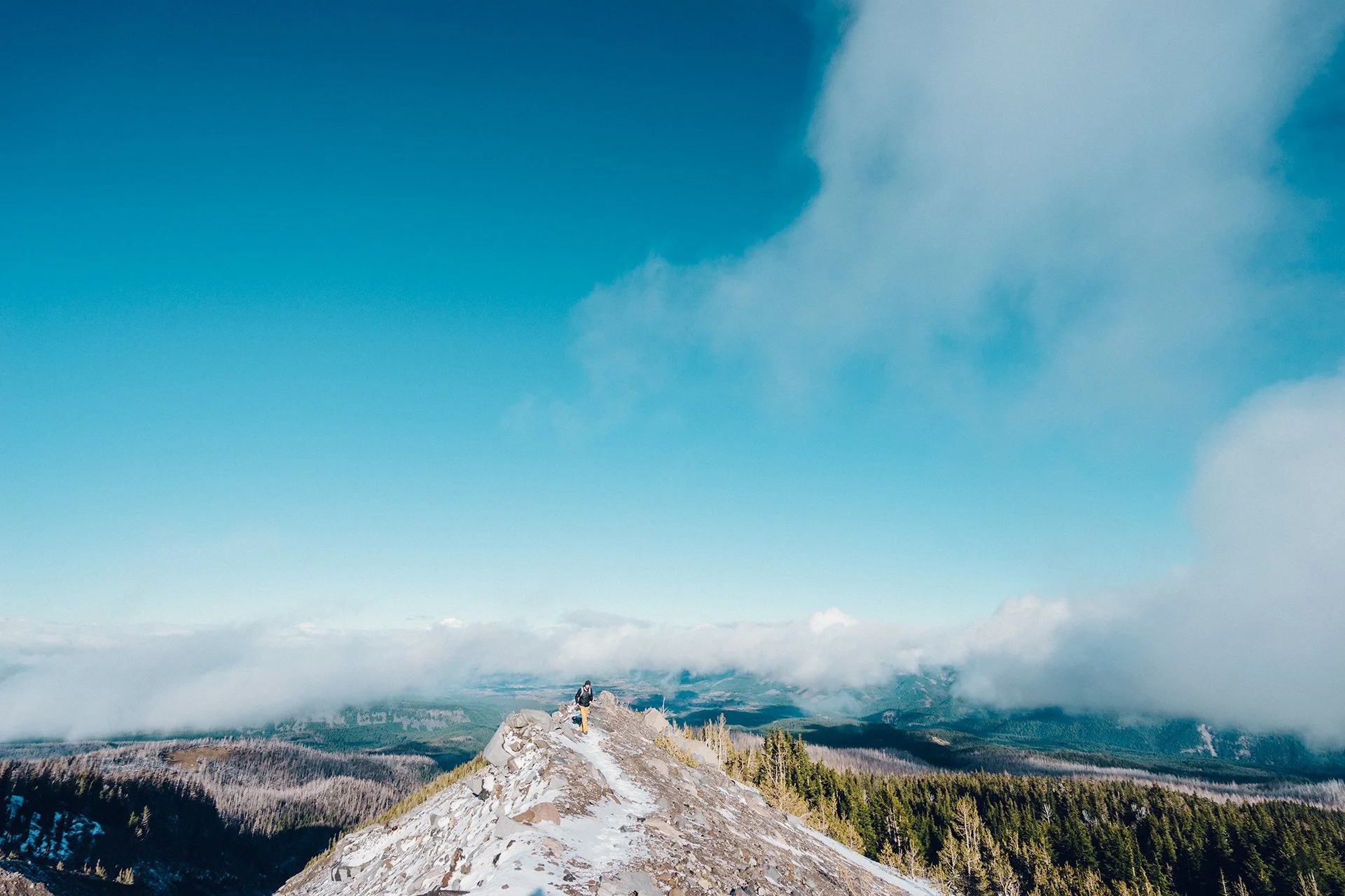 Mountain ridge with snow, person hiking, view of forest and clouds, blue sky.