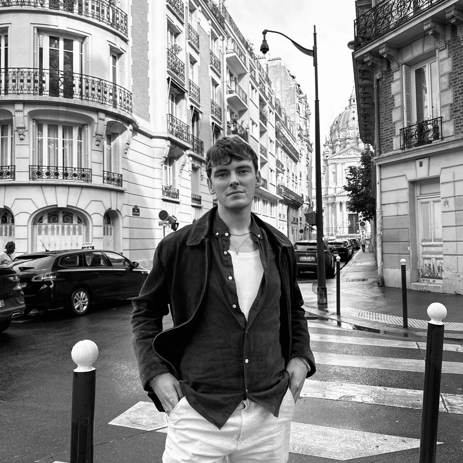 A young man standing on a city street corner, wearing a dark jacket and light-colored pants, with buildings, parked cars, and a church dome in the background.
