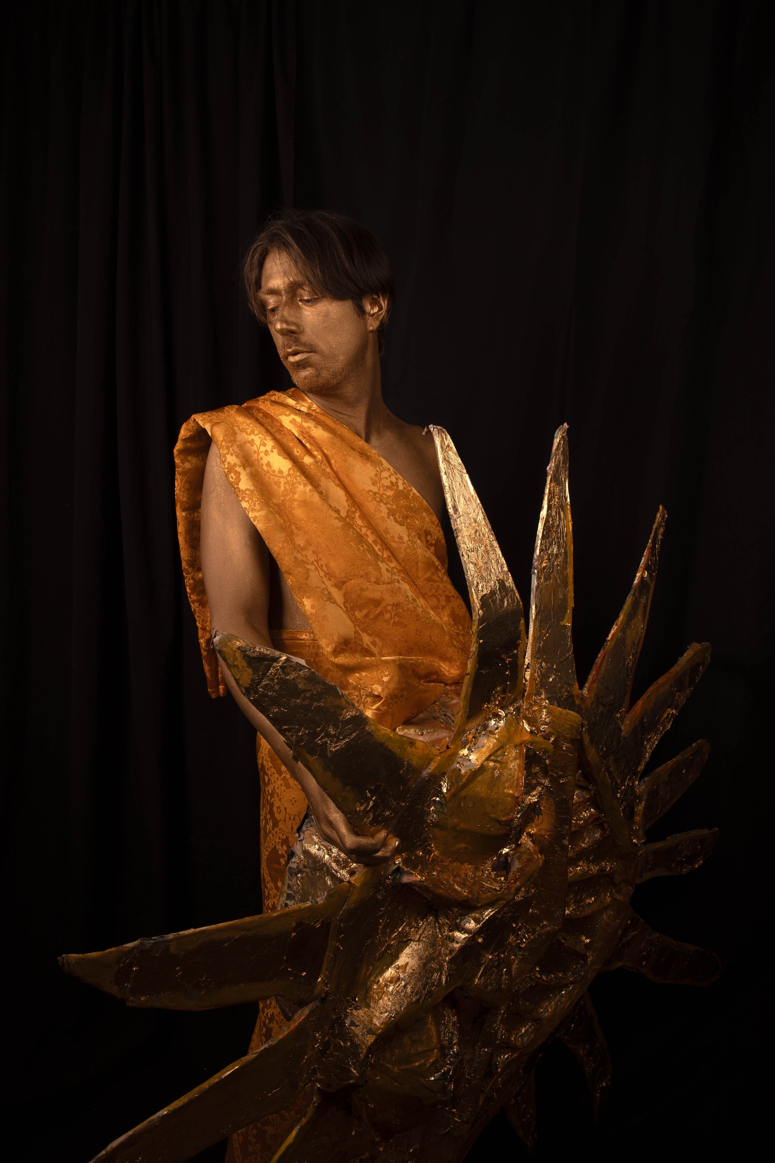 Glasgow musician Peter Cat dressed in a gold patterned fabric holding a gold and black spiked sculpture, standing against a dark background.
