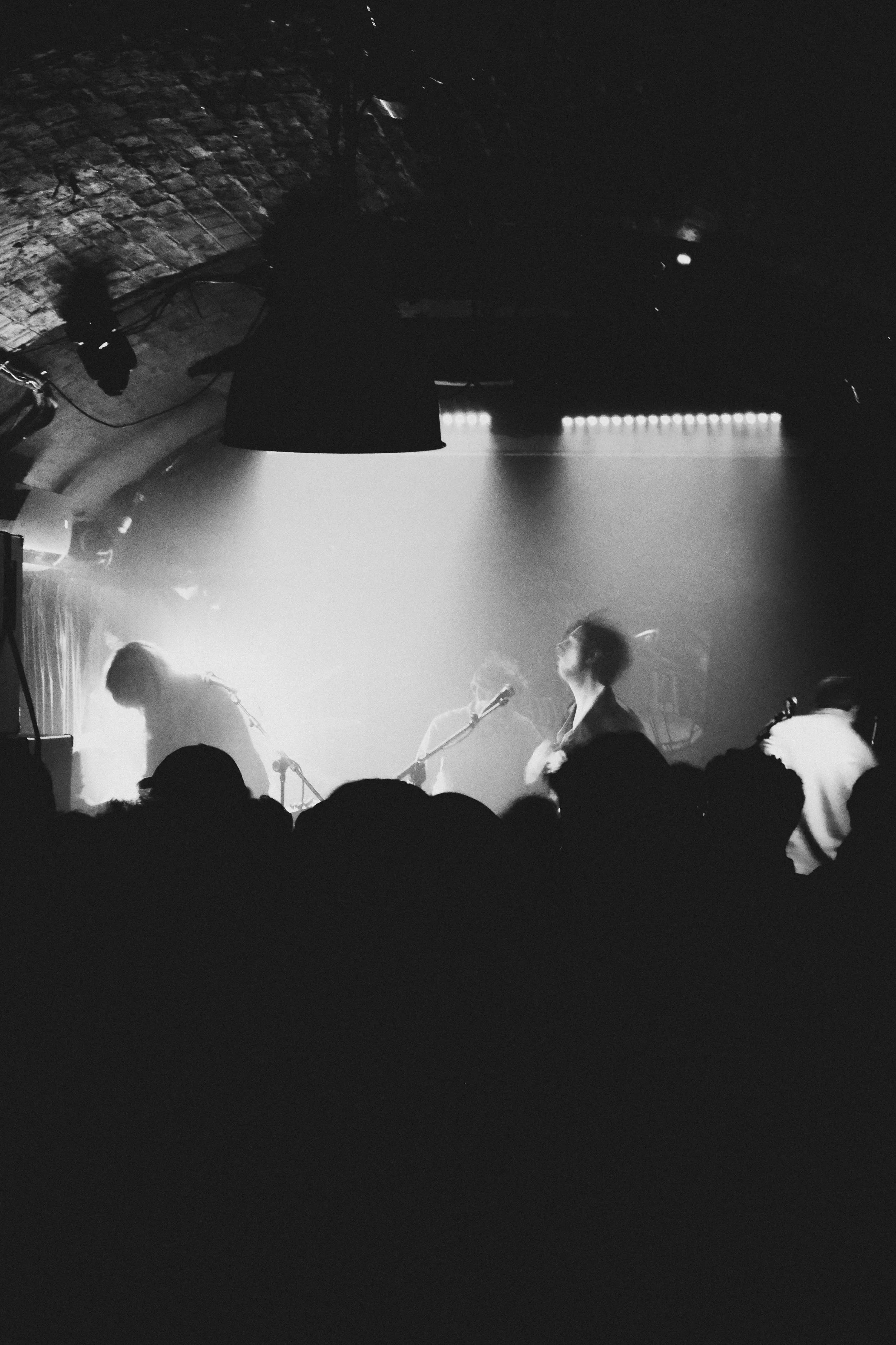 Black and white photo of the Glasgow band Peter Cat performing on stage in a dimly lit venue, with silhouettes of audience members in the foreground.