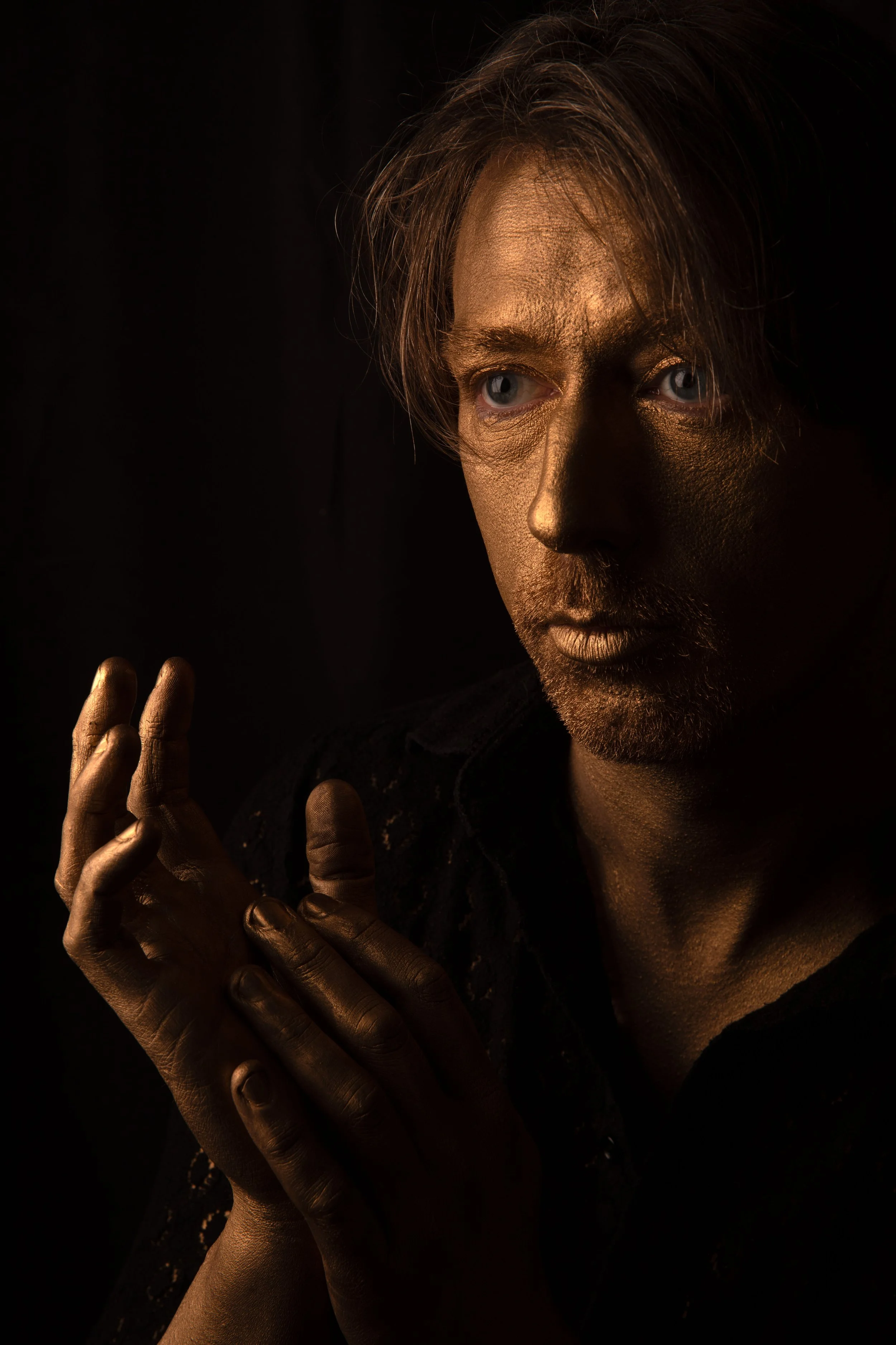 Glasgow musician Peter Cat with gold painted skin and face, looking to the side, holding hands together against a dark background.