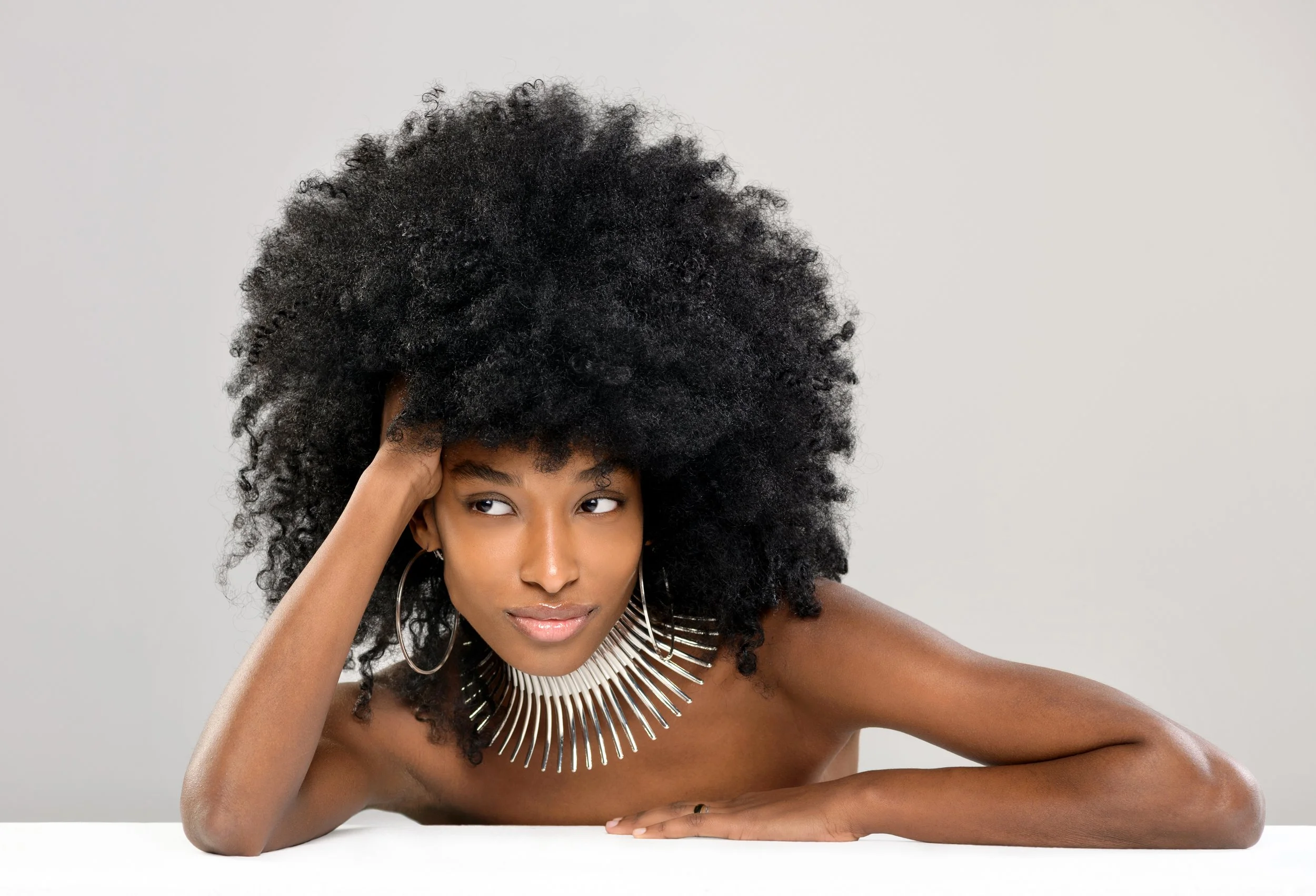 Young woman with large curly black hair lying on her side, resting her head on her hand, wearing a statement necklace and hoop earrings, against a plain light background.