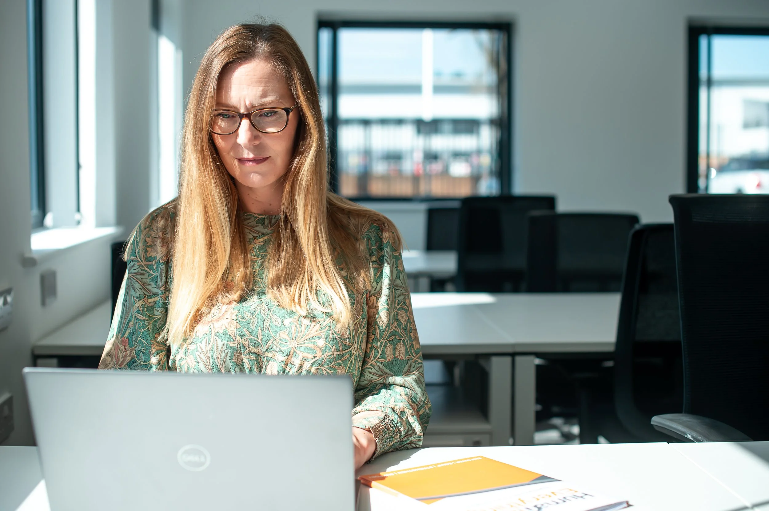 A woman working on a laptop in an office with large windows and empty chairs.