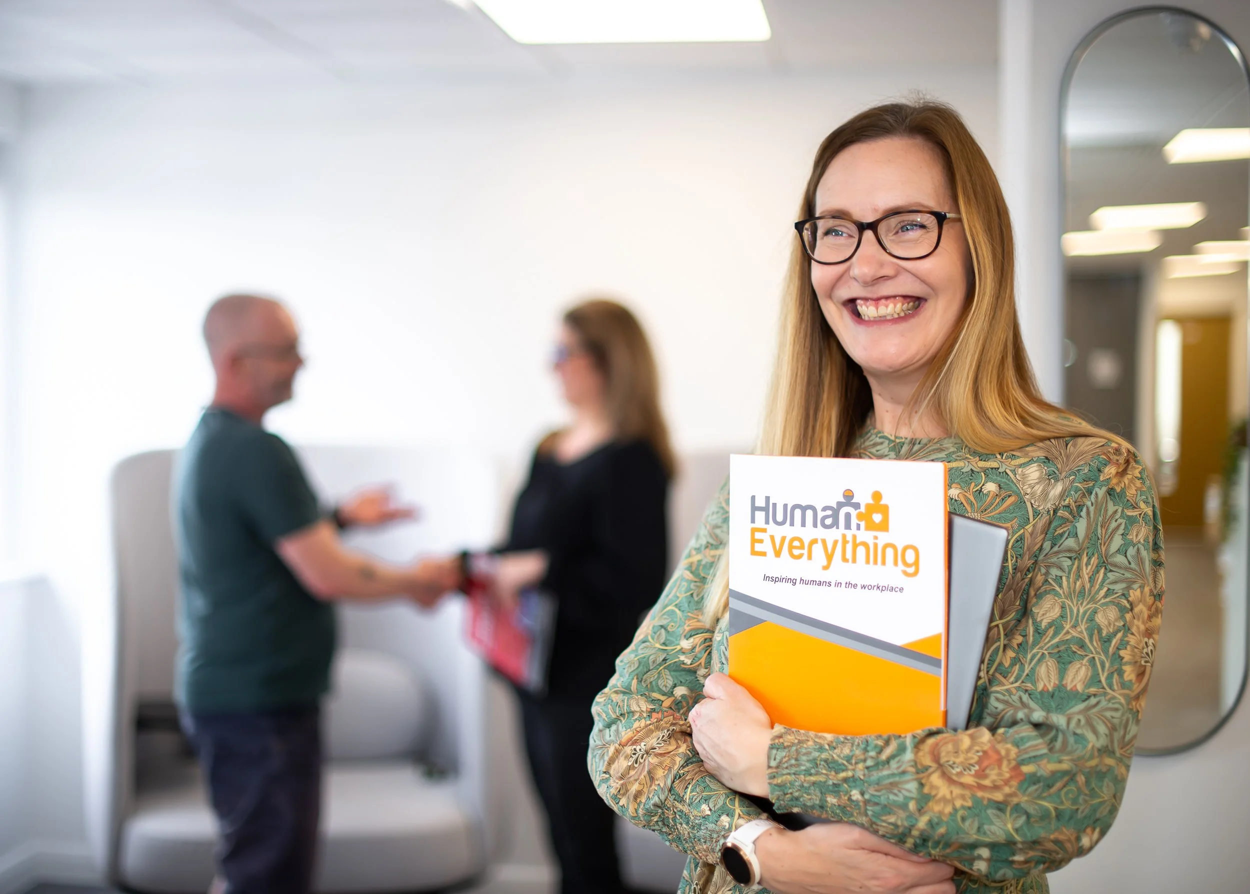 A smiling woman with glasses holding a folder that reads 'Human Everything' in an office setting. In the background, two blurred people are shaking hands.