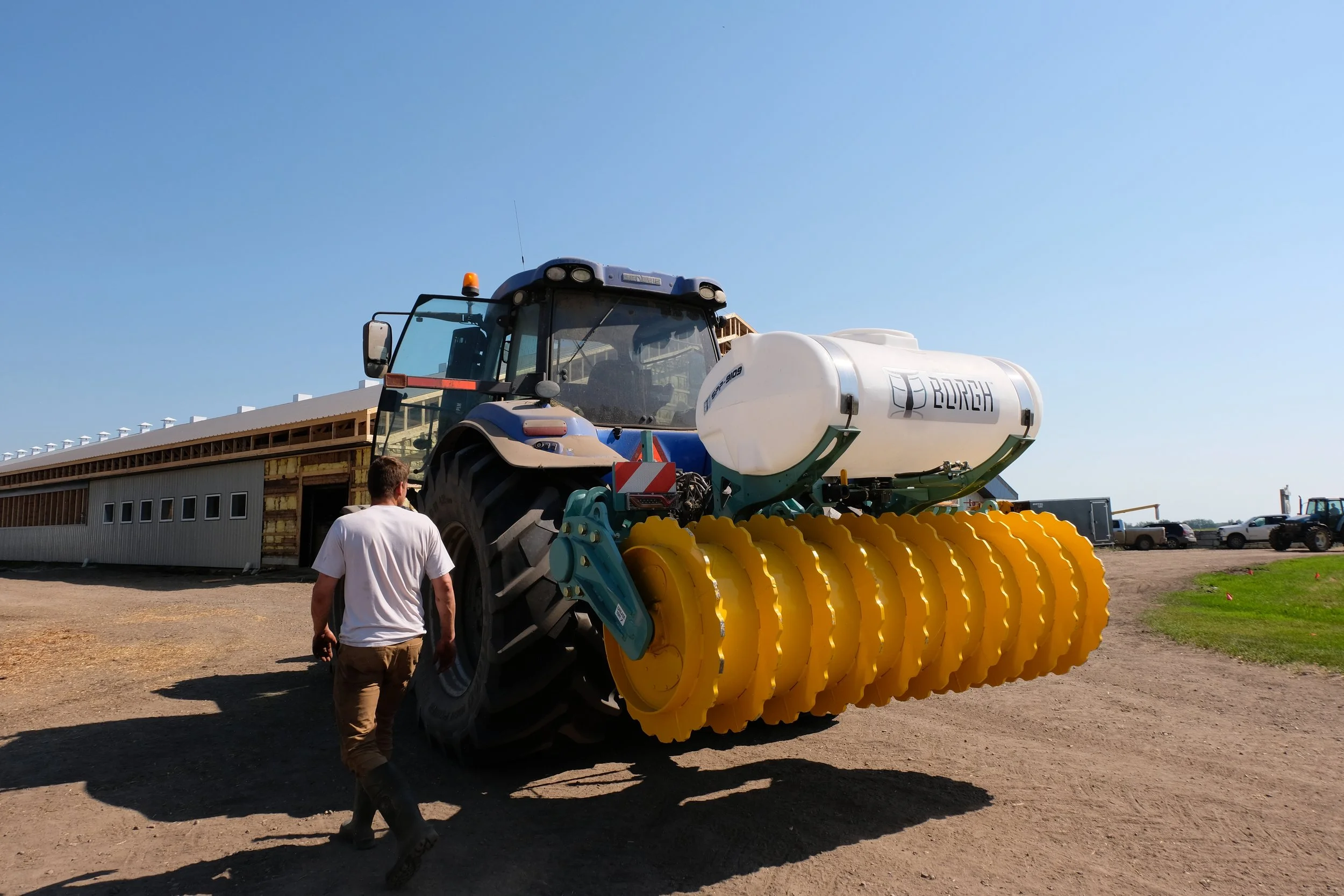 A dairy farmer getting into New Holland tractor that has a silage pile packer attachment connected to it's 3-point hitch. There is also a white 200 gallon inoculant tank on the silage pile packer.