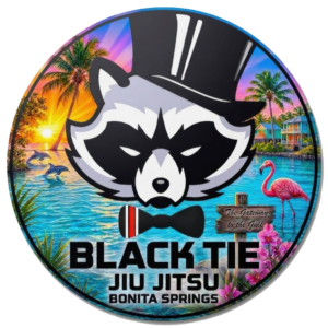 A colorful design featuring a raccoon wearing a top hat and bow tie, with a sunset, palm trees, flamingos, and water in the background, and the text 'Black Tie Jiu Jitsu Bonita Springs'.