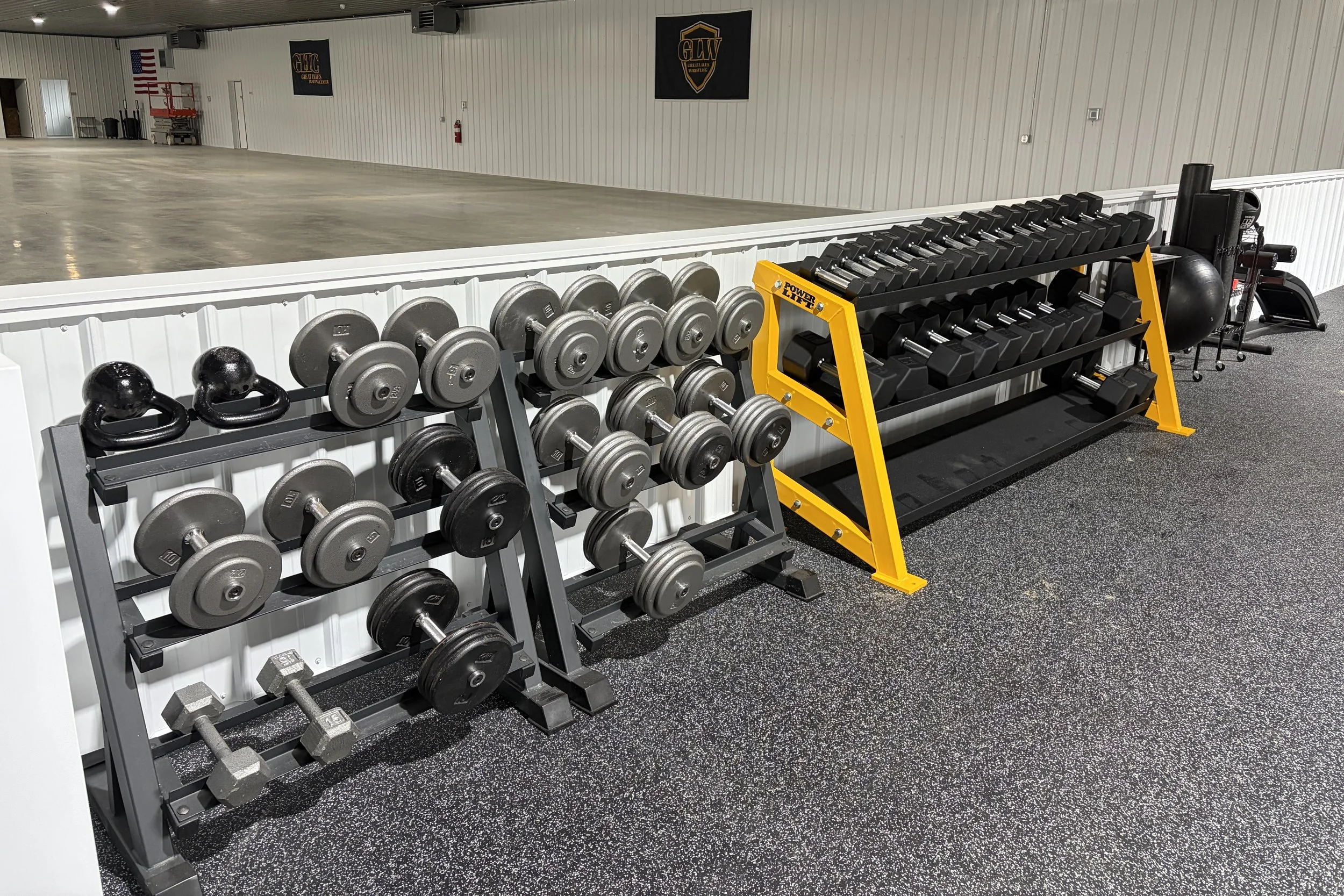 A gym with racks of dumbbells, kettlebells, and a yellow Power Lift squat rack. In the background, there are banners on the wall and a fire extinguisher. The gym has a polished concrete floor and cream-colored corrugated metal walls.