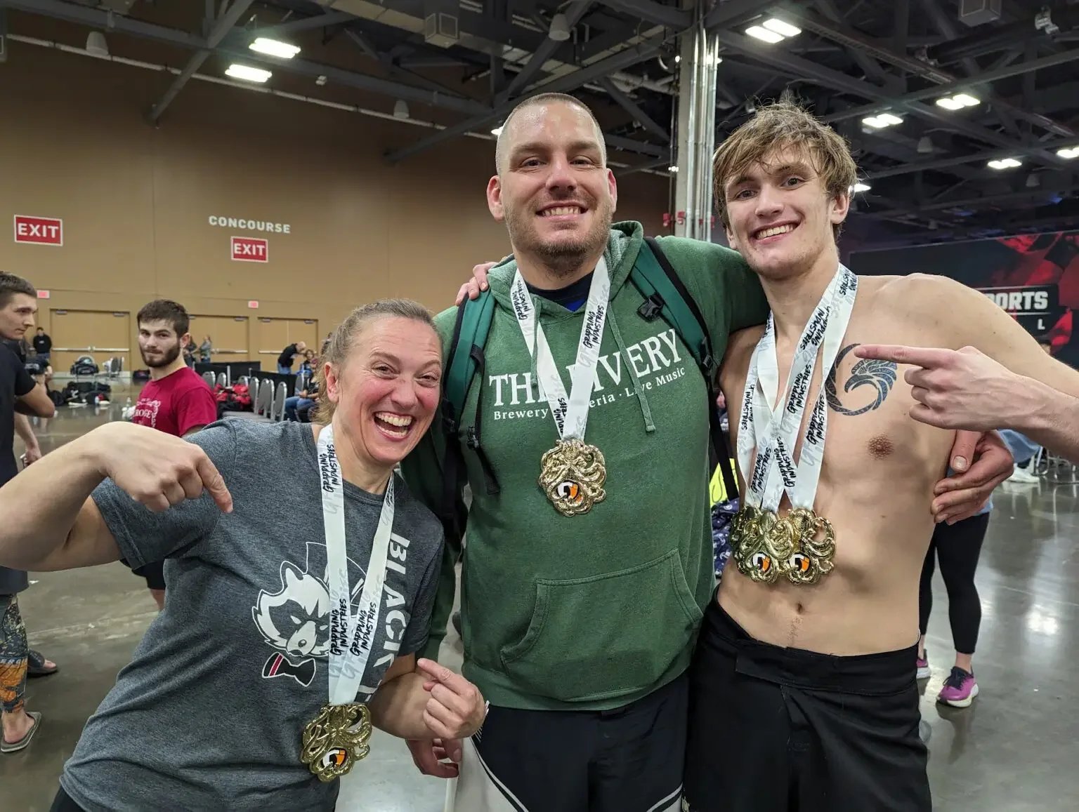 Three individuals with medals around their necks celebrating at a sports event, smiling and pointing to their medals. The person on the right is shirtless and has a tattoo, the person on the left is wearing a gray shirt, and the person in the middle is wearing a green hoodie.