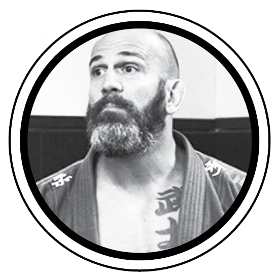 Black and white photo of a man with a beard and shaved head, wearing a martial arts gi with a tattoo on his chest, standing indoors.