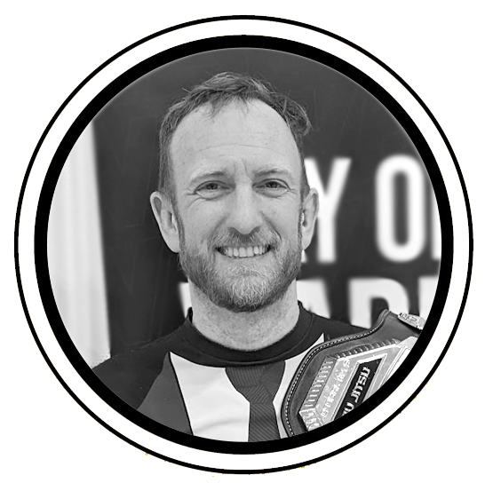 Black and white photo of a smiling man with short hair and a beard, wearing a sports jersey, holding a championship belt.