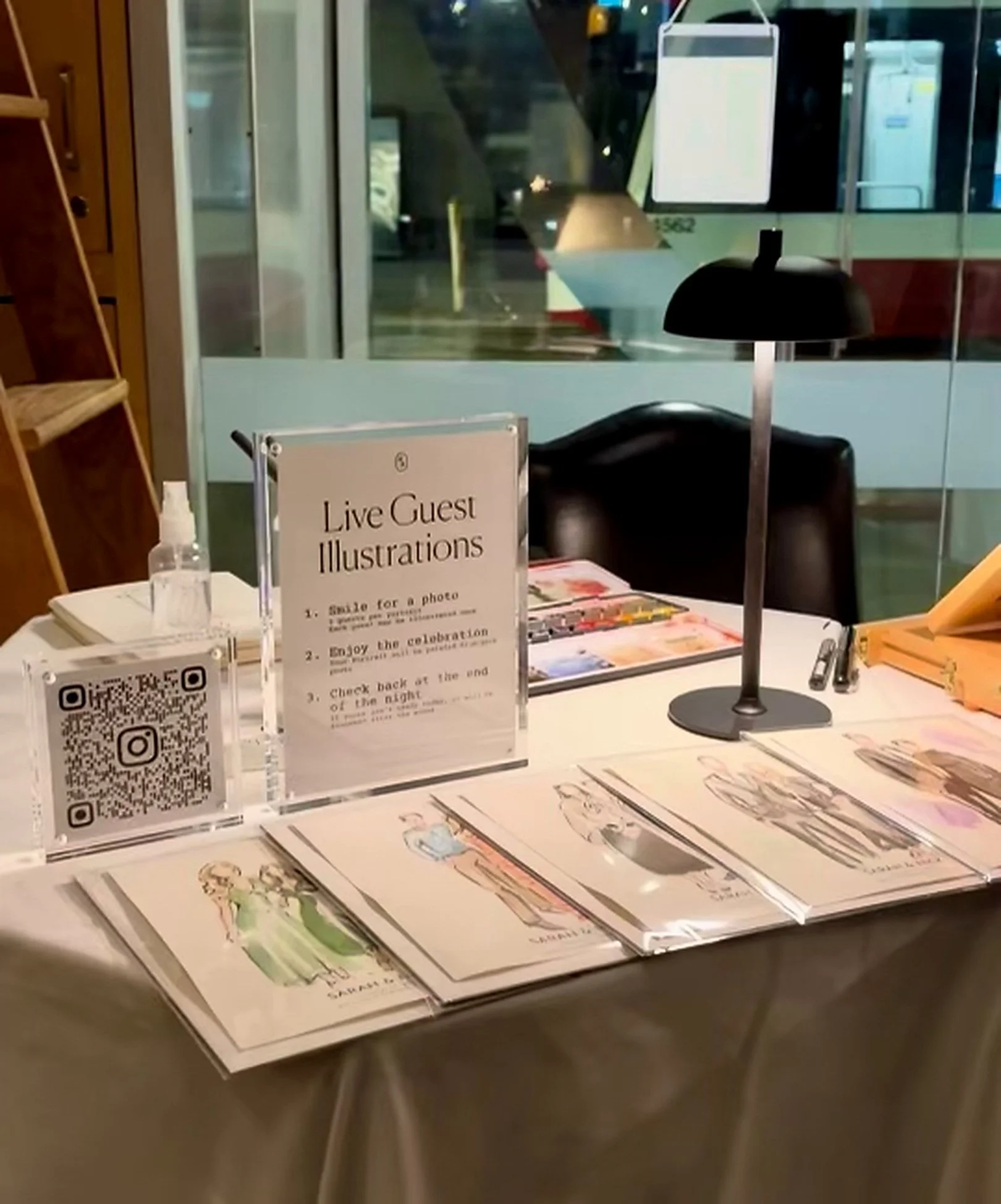 Table displaying illustrated guest portraits and artwork with a sign about live guest illustrations, QR code, and art materials, positioned near a window and a hand sanitizer bottle.