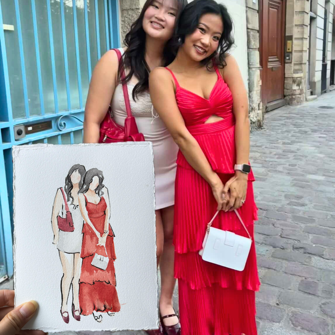 Two women standing on a cobblestone street, smiling and posing for a photo. One is wearing a white dress with pink accessories, the other is in a red dress. Someone is holding a piece of fabric with a fashion sketch of two women, one in a white dress