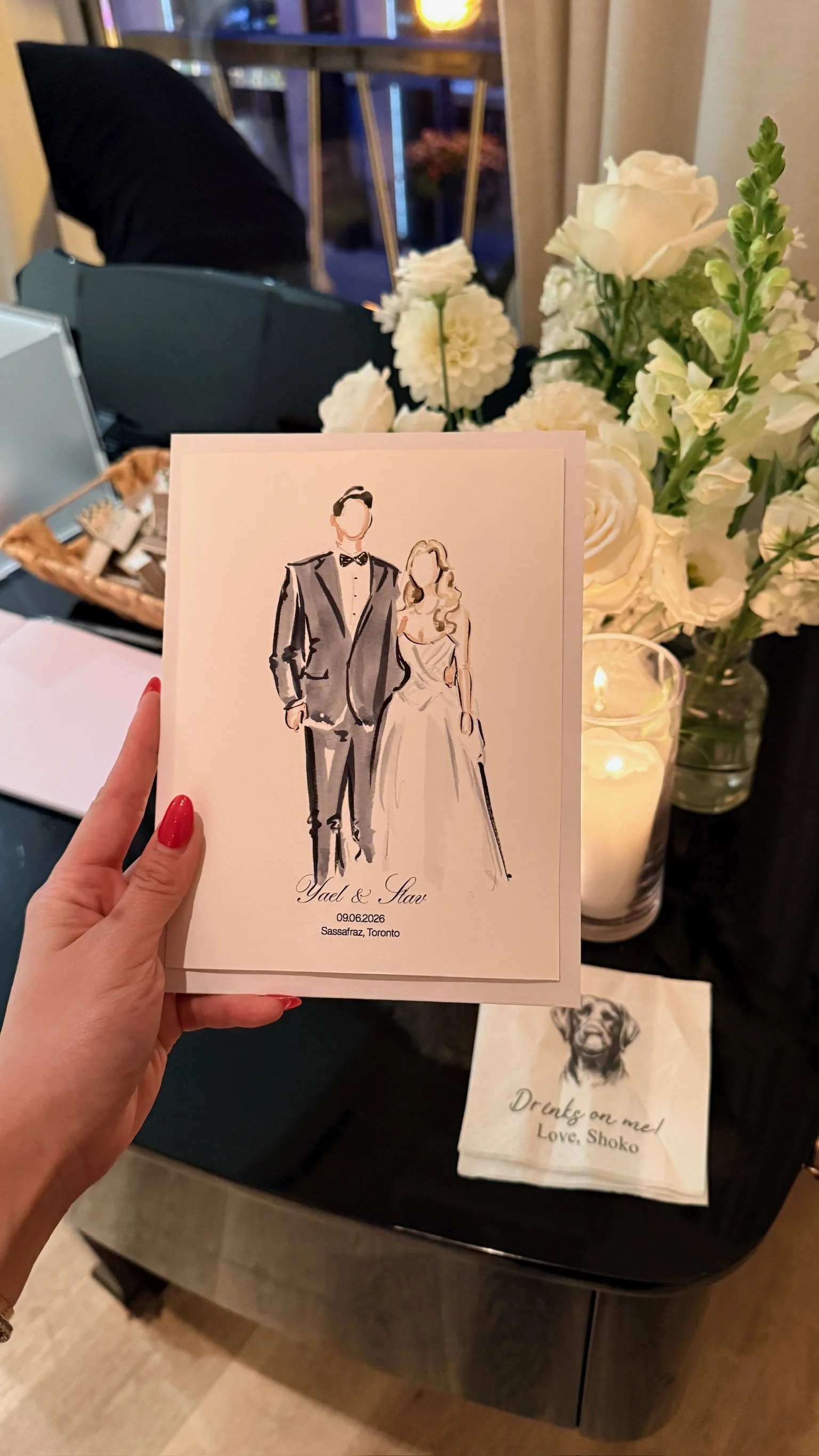 A hand holding a wedding card illustrating a couple in formal attire, with the names 'Yael & Stav' and the date '09.06.2026' at Sasafraz, Toronto. The background features white flowers and a lit candle.