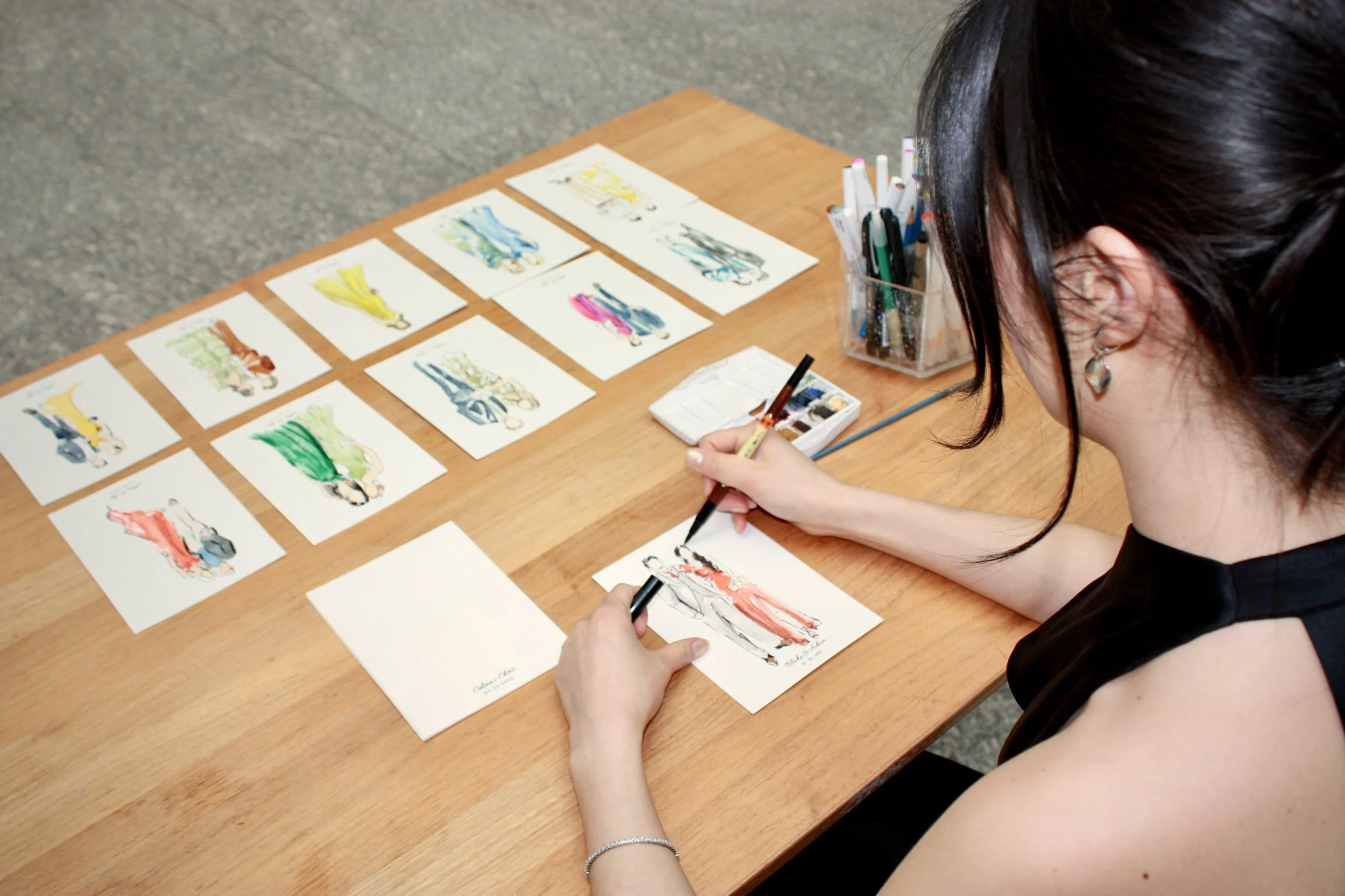 A woman is painting fashion sketches of women in colorful dresses on small pieces of paper at a wooden table, with similar sketches arranged in rows in front of her.
