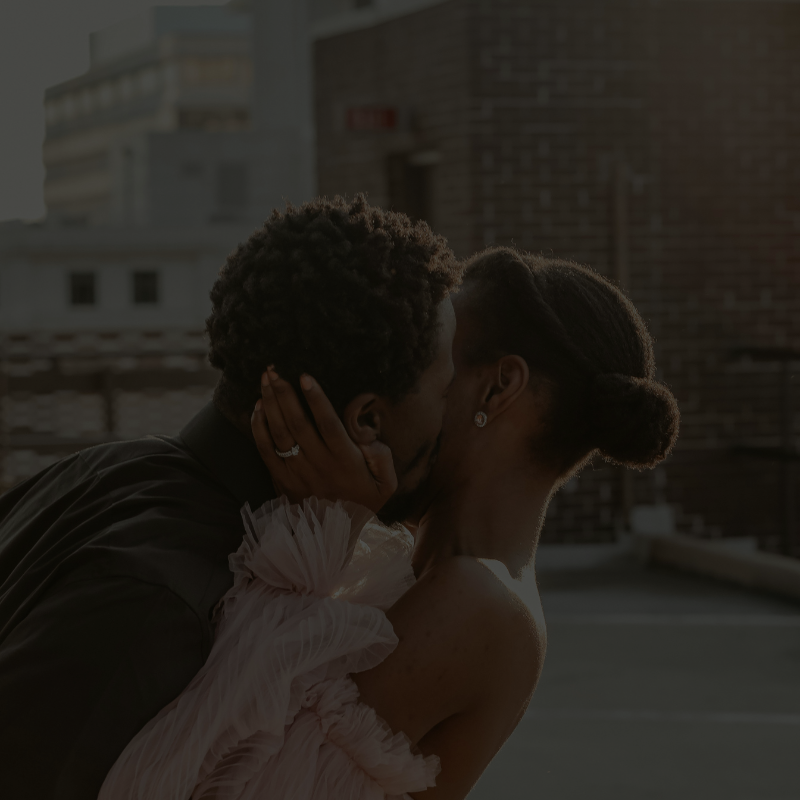 Affluent Black couple sharing an intimate kiss on a rooftop at sunset, dressed in formal evening wear.