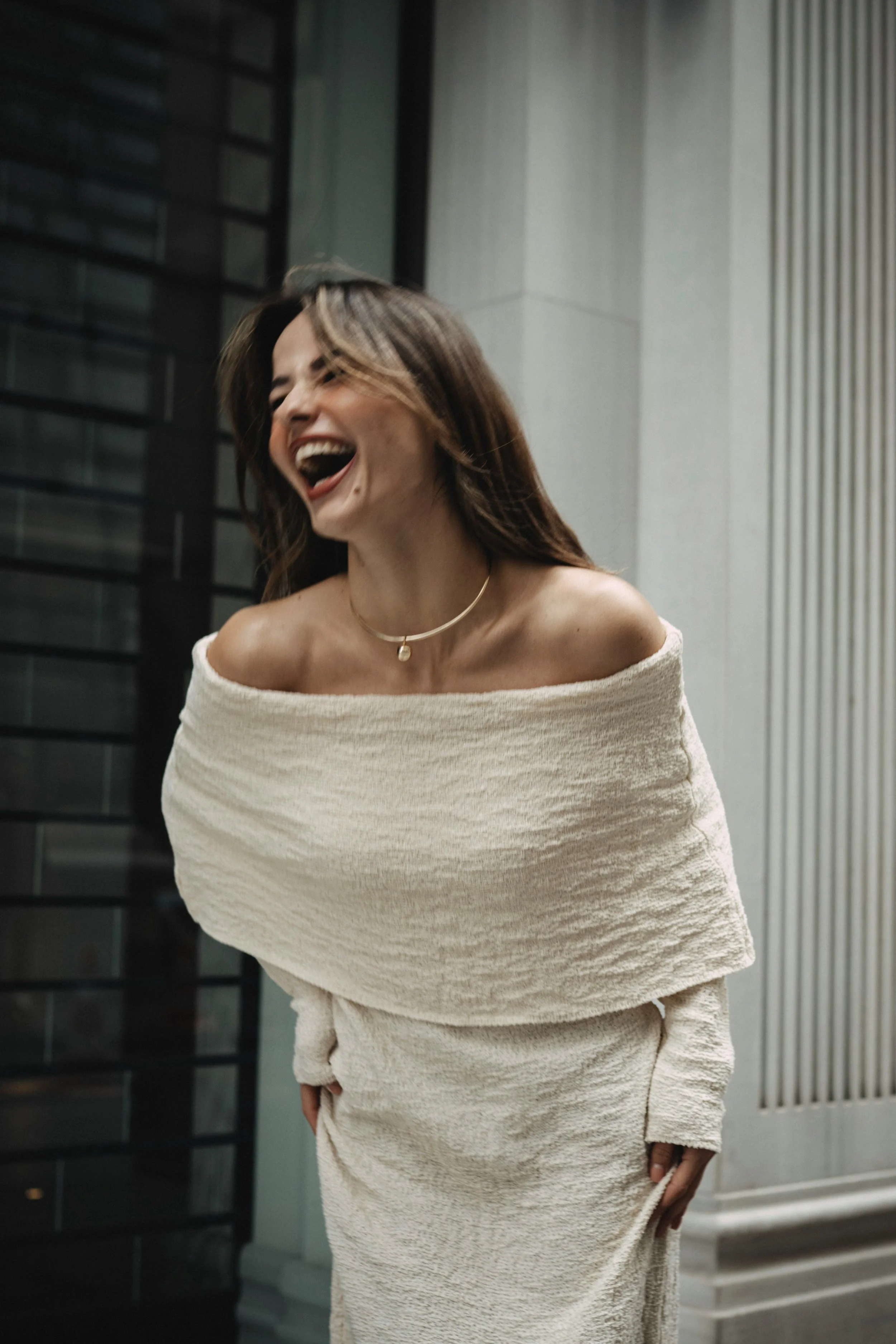 Woman laughing confidently in an off-the-shoulder white dress, standing in front of a modern building.