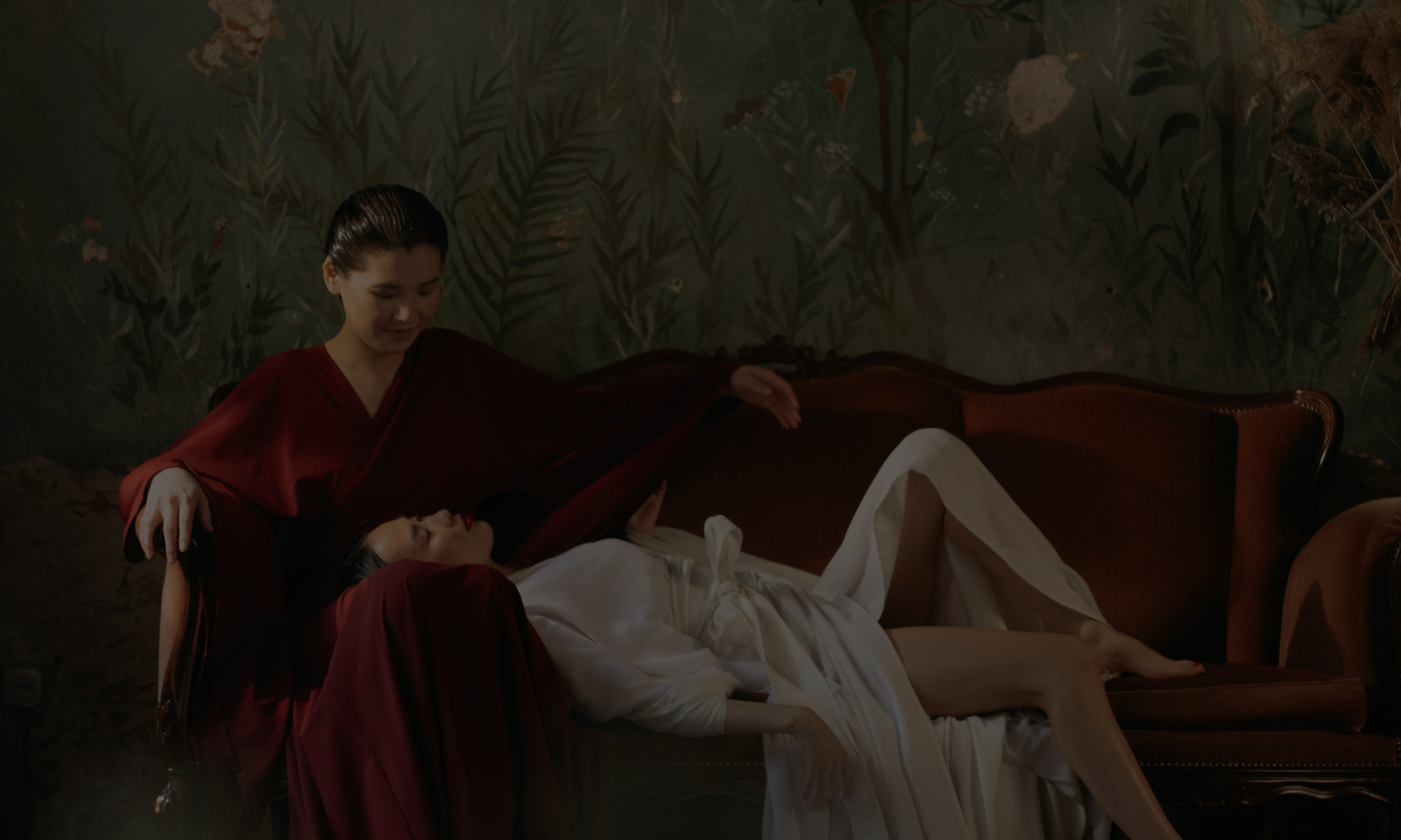 Queer Asian couple sharing a quiet, intimate moment on a vintage couch, with one partner resting in the other's lap in soft, moody lighting.