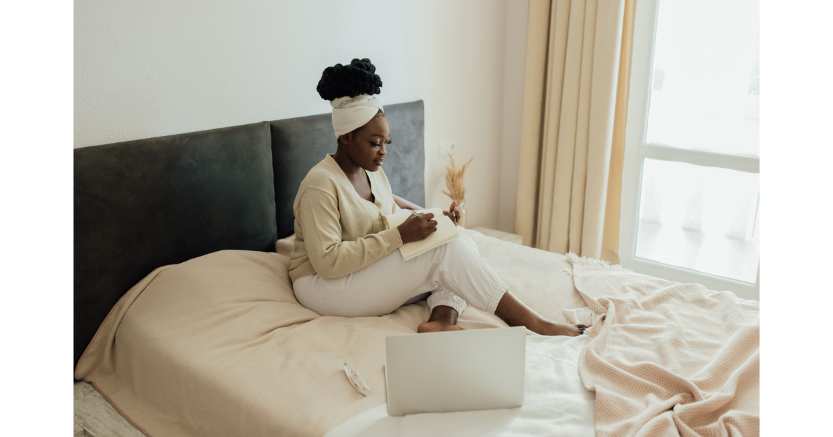 Black woman journaling in a notebook while sitting comfortably on a bed, with a laptop open nearby in a softly lit bedroom.