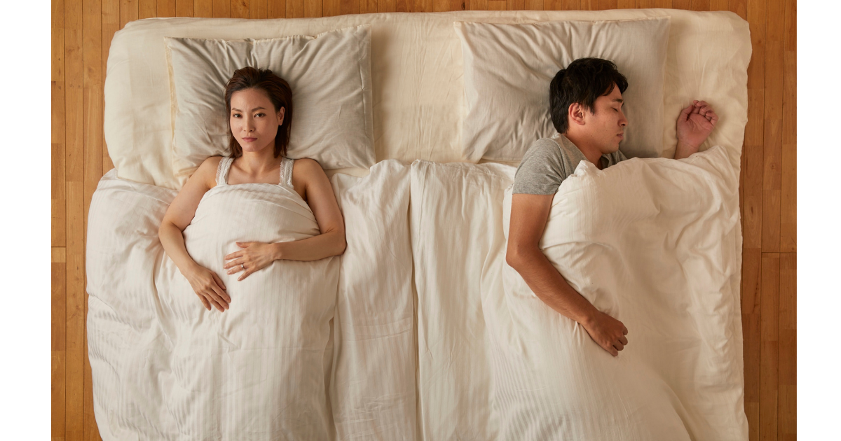 Woman awake and man asleep while lying back to back in bed, representing emotional distance or relationship tension.