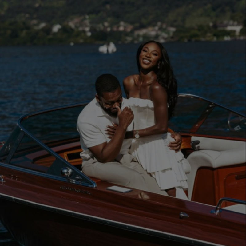Joyful Black couple laughing together on a luxury speedboat with scenic water and mountain views in the background.