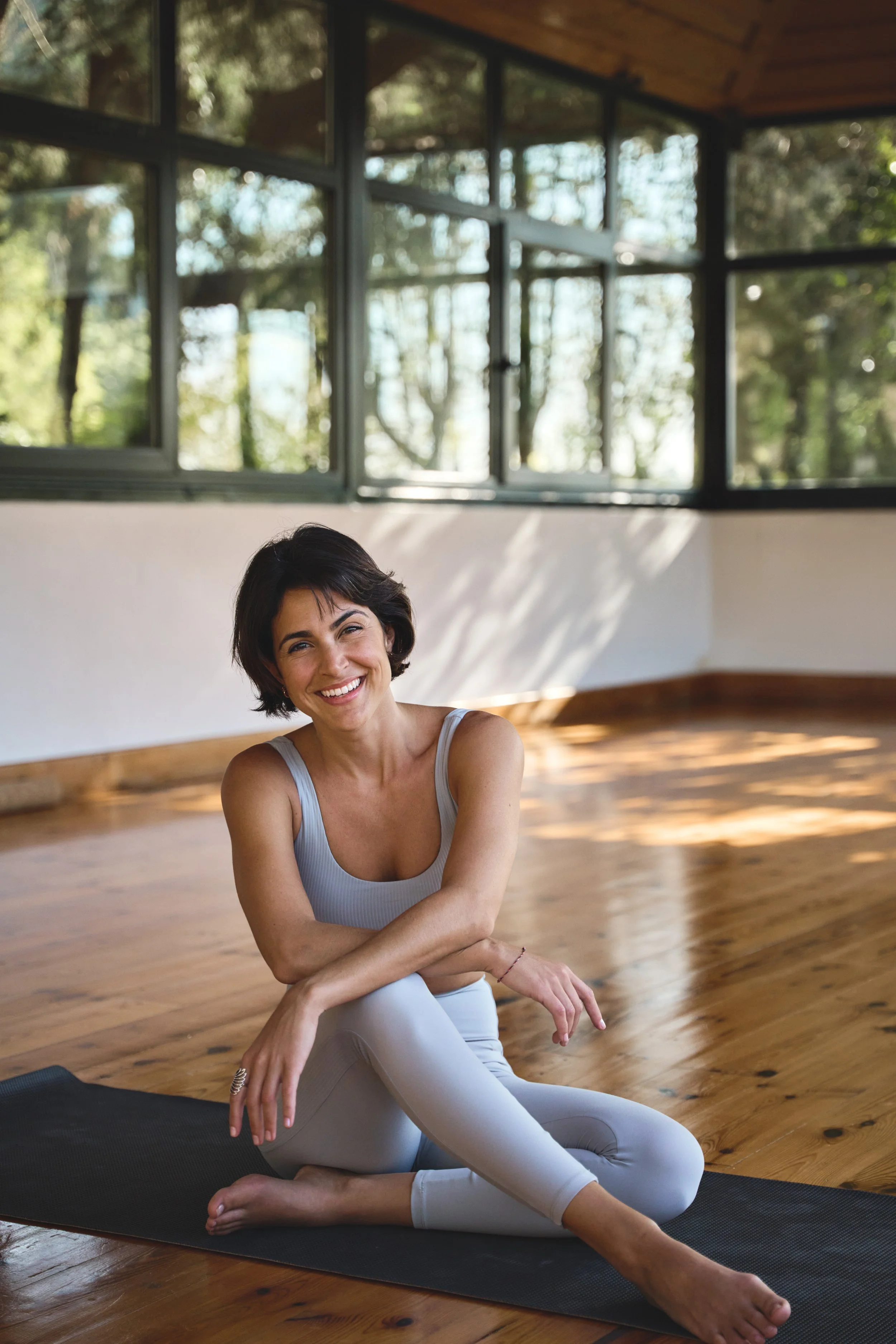 Woman smiling, sitting cross-legged on a yoga mat in a sunlit room with large windows.