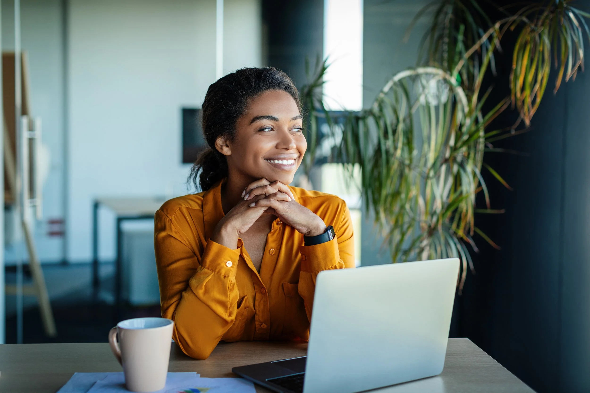 Smiling woman in mustard blouse sitting at desk with laptop and coffee cup in a modern office with plants.