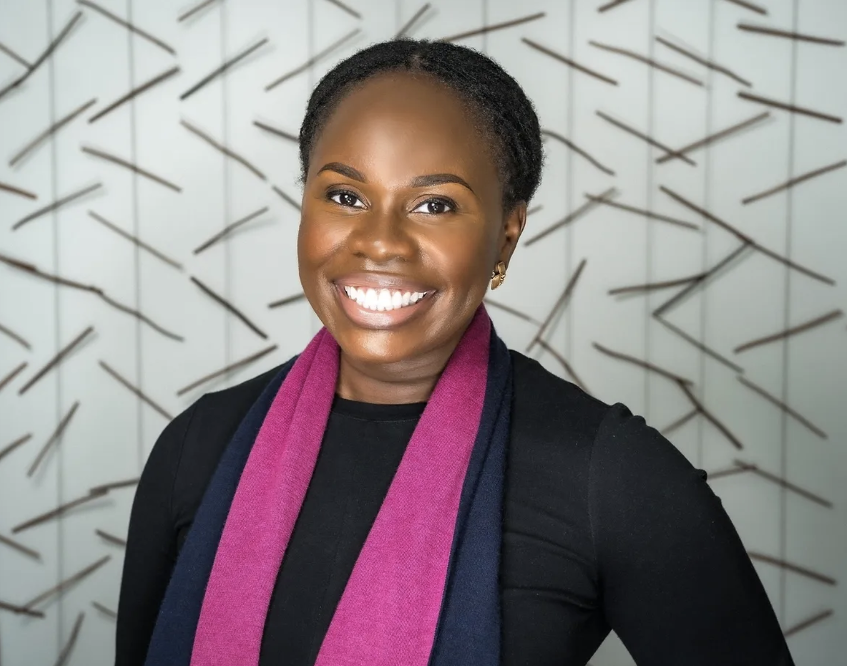 Smiling person with short hair wearing a black top and purple scarf, standing before a patterned background.
