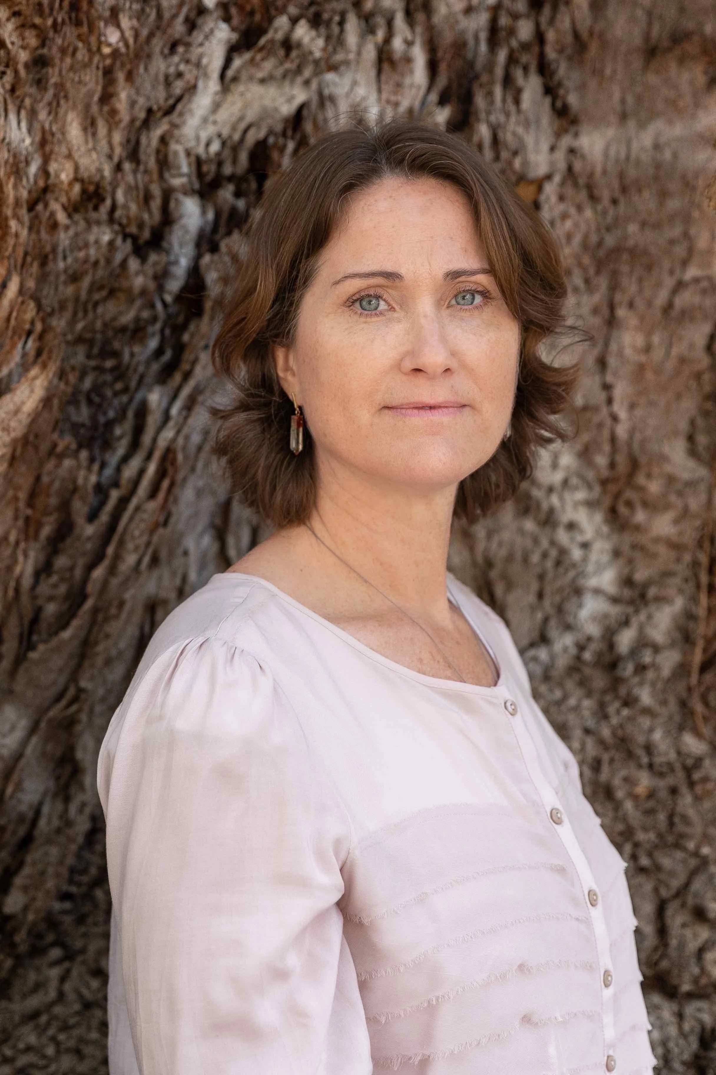 A person with short brown hair and a light shirt standing in front of a textured tree bark background.