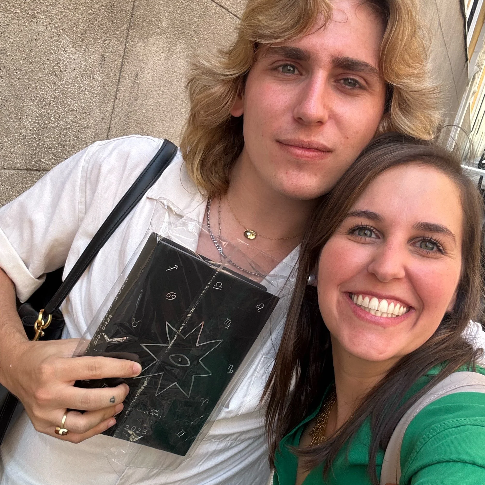 Dos personas sonriendo y tomándose una selfie, una mujer con cabello castaño oscuro y camiseta verde, y un hombre con cabello rubio largo y camiseta blanca, sosteniendo una caja con un diseño de sol y letras en inglés.