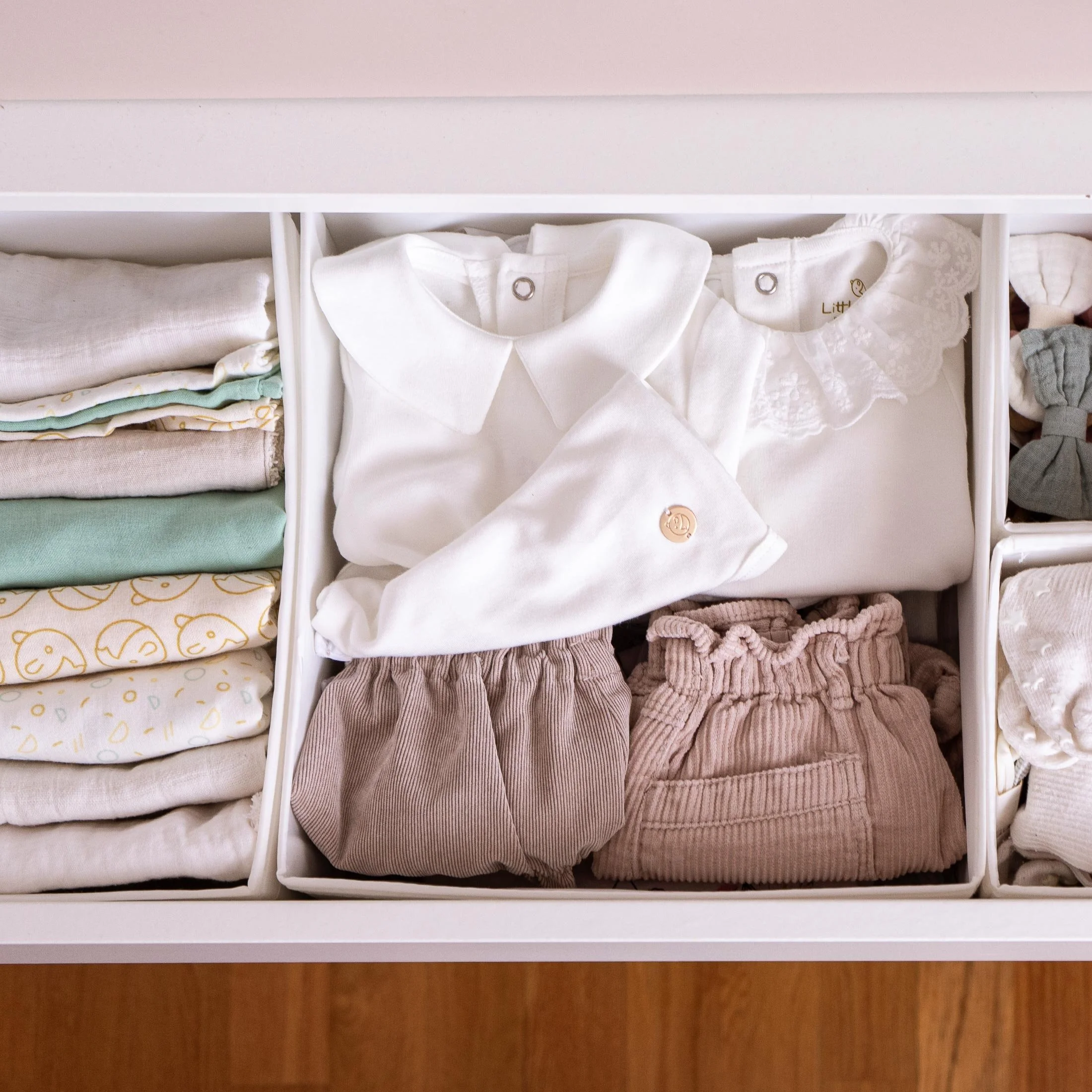 Organizador con ropa de bebé, incluyendo un conjunto con camisa, pantalones cortos, y otros accesorios en tonos neutros y pastel.