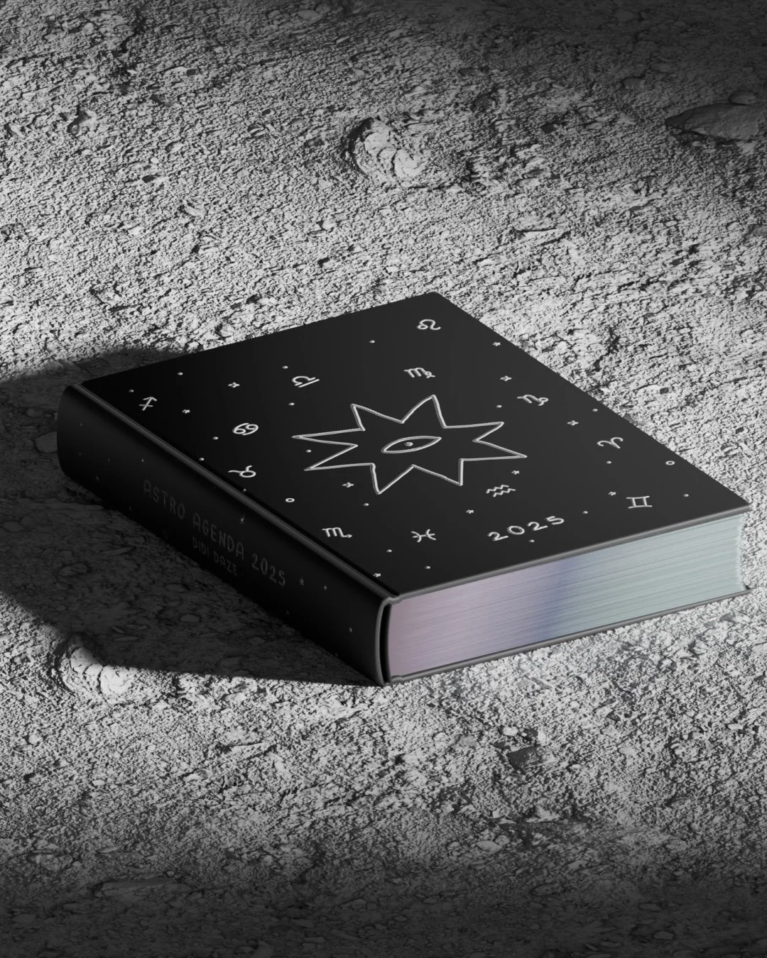 Agenda astral 2025 de color negro con ilustración de un ojo y un sol en la portada, en un ambiente con superficie de tierra.