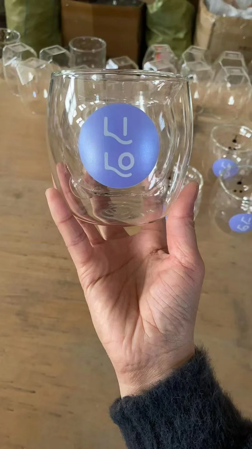 Vaso de vidrio con etiqueta azul que dice 'LUV', sostenido por una mano en un entorno con otros vasos y etiquetas similares en el fondo.