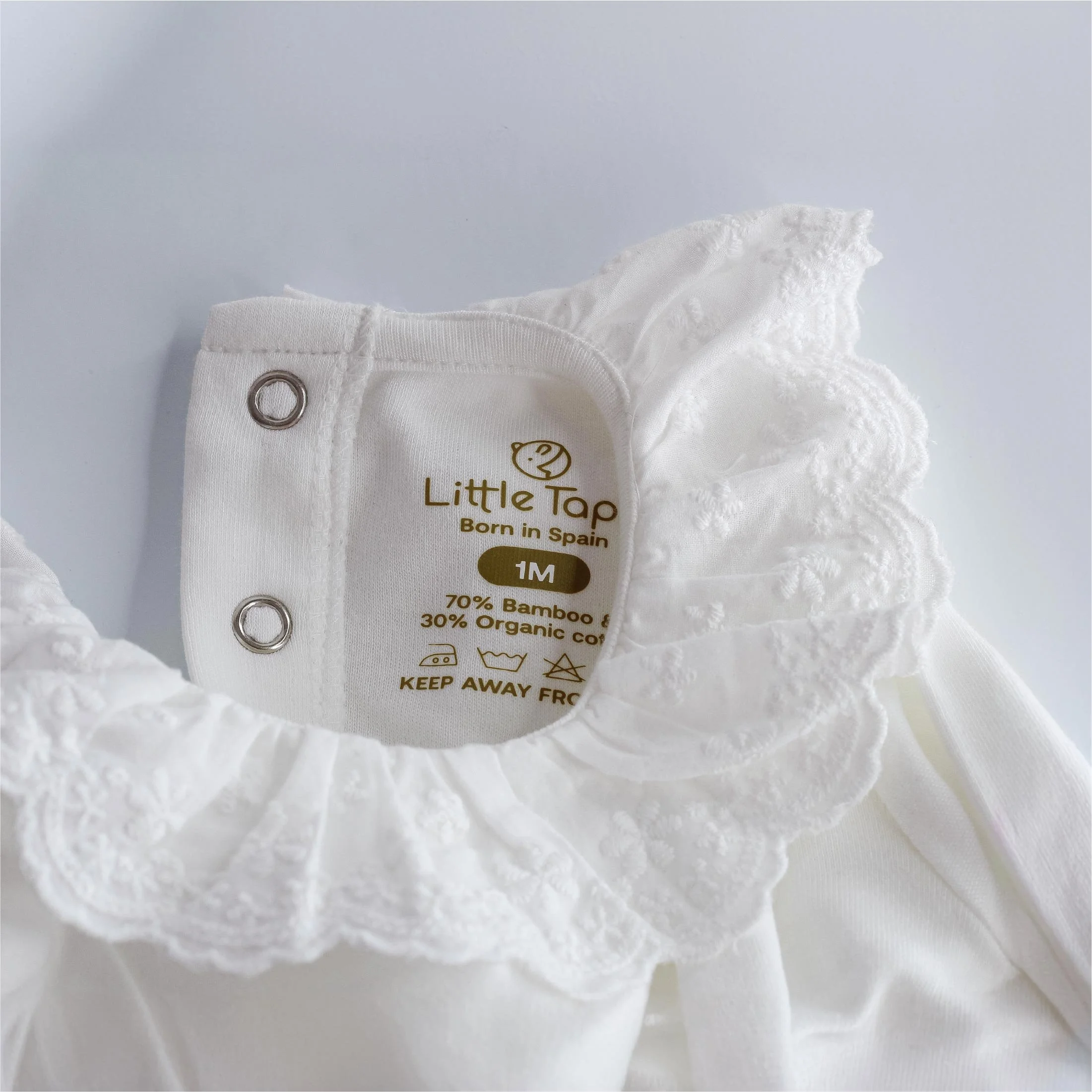 Etiqueta de ropa para bebé blanca con detalles de encaje, marca Little Tap, indica talla 1 mes, 70% bambú, 30% algodón orgánico, cuidados de lavado y símbolos de precaución.