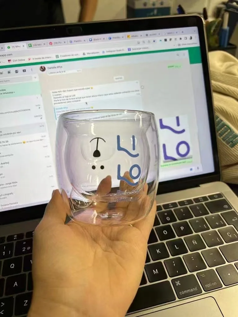 Una mano sostiene un vaso de cristal con dibujos de una cara de payaso y letras azules, frente a una computadora portátil con la pantalla visible.
