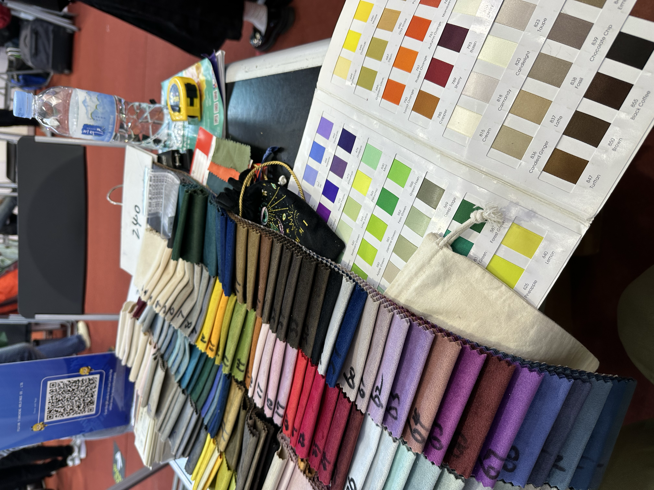Muestras de tela de colores variados organizadas en filas, junto a muestras de pintura en una caja con un diagrama de colores. También se observan objetos como una botella de agua, una cinta métrica, papeles y bolsas pequeñas, en una mesa en un ambie