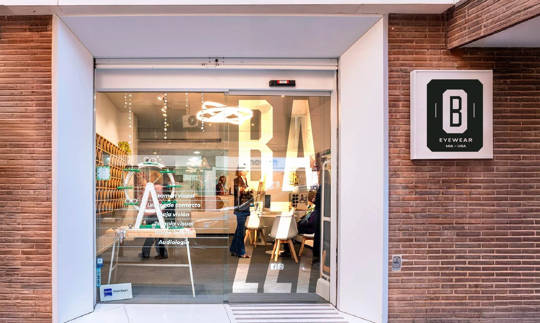 Vista de la entrada de una tienda de gafas con cristalera grande, en una calle de ladrillo, con personal y clientes en el interior y cartel con texto en la cristalería.
